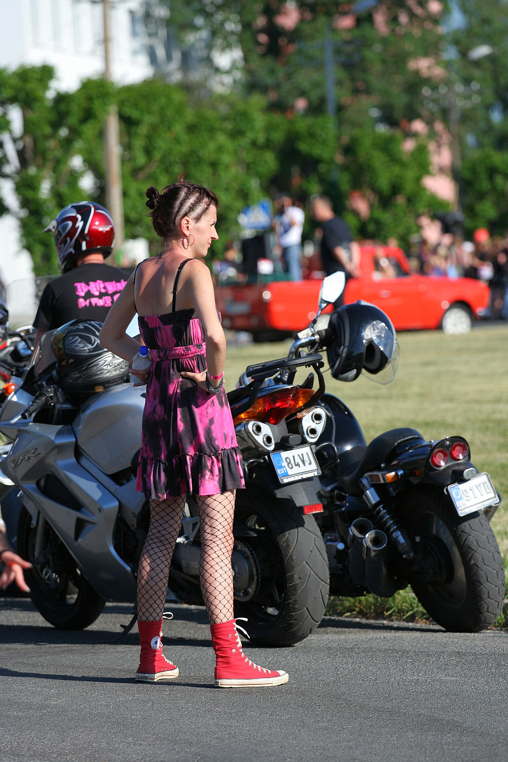 A person with braided hair, wearing a pink and black sleeveless dress, fishnet stockings, and red high-top sneakers, stands in front of a row of motorcycles. Another person in a helmet and black shirt sits on one of the bikes. The scene has a punk vibe with trees and a red car in the background, evoking Laulupidu energy.
