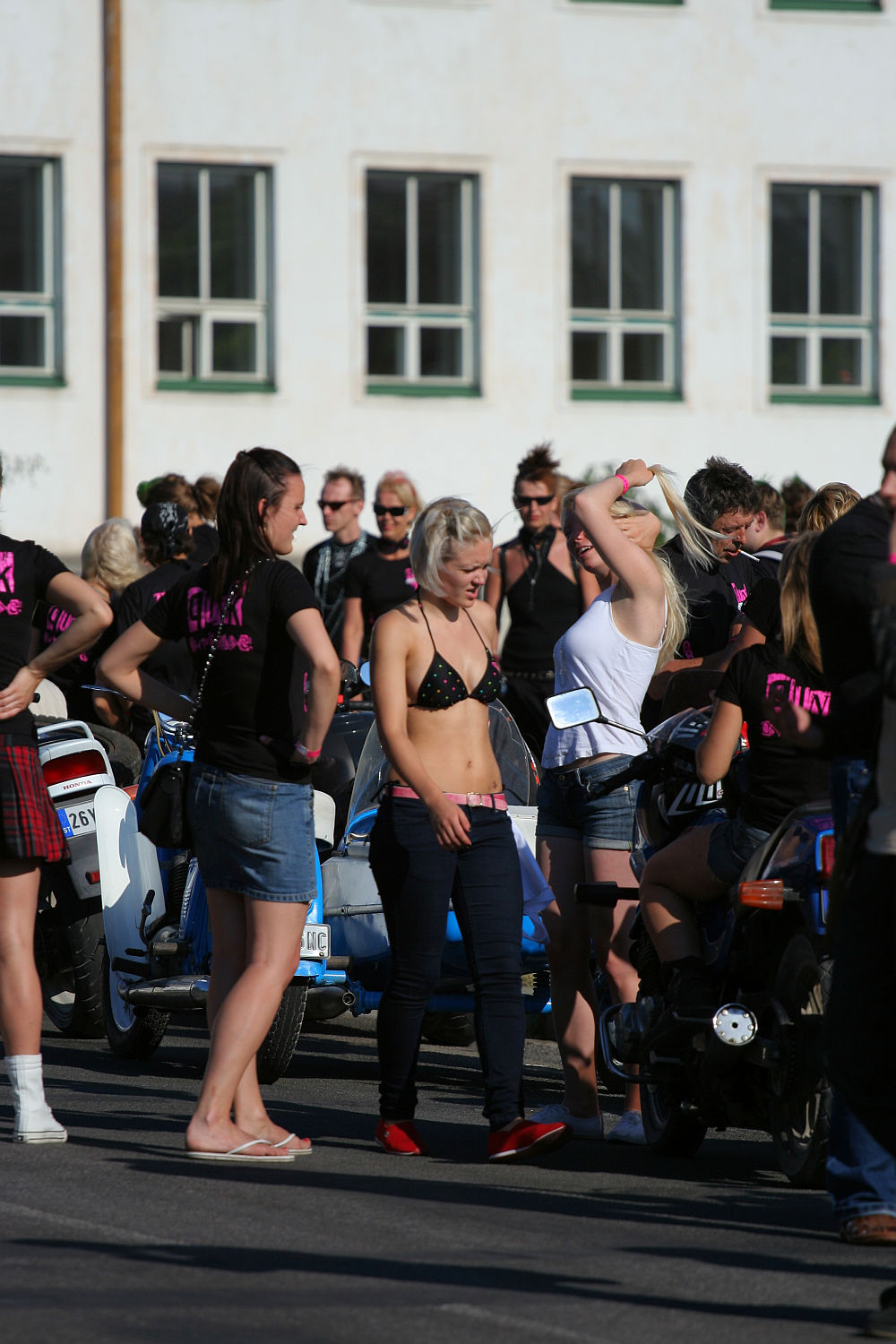 A group of people gather at an outdoor event, standing near motorcycles. The central person wears a black bikini top and red shoes, giving off a distinct punk vibe. Another person on the right adjusts their white top. Many attendees wear black shirts with pink writing. The background features a white building with windows, reminiscent of scenes from Laulupidu celebrations.