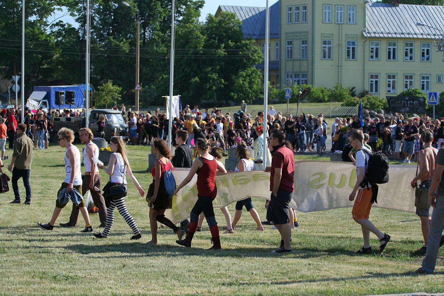 A group of people walk through a grassy field holding a large, partially visible banner. The individuals, dressed in casual summer clothing including shorts, tank tops, and boots with a hint of punk style, seem energized. In the background, a large crowd gathers reminiscent of Laulupidu celebrations, with trees and buildings visible farther away.