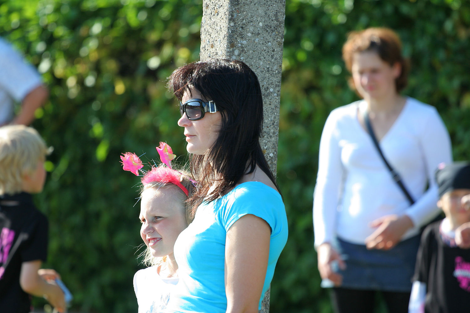 A woman in sunglasses and a blue shirt stands beside a young girl wearing a headband with pink decorations. They are outdoors, leaning against a tree with lush greenery in the background. Other people, possibly gathering for the vibrant Laulupidu festival, appear indistinctly in the background.