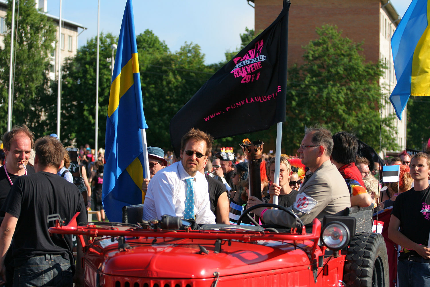 A colorful parade weaves through the streets, with some participants riding a red vehicle. Two prominent figures, one in a white shirt and blue tie, and another in a beige suit, wave flags. Black and Swedish flags flutter alongside an onlooker's small Estonian flag. Trees and buildings frame this festive Laulupidu scene.
