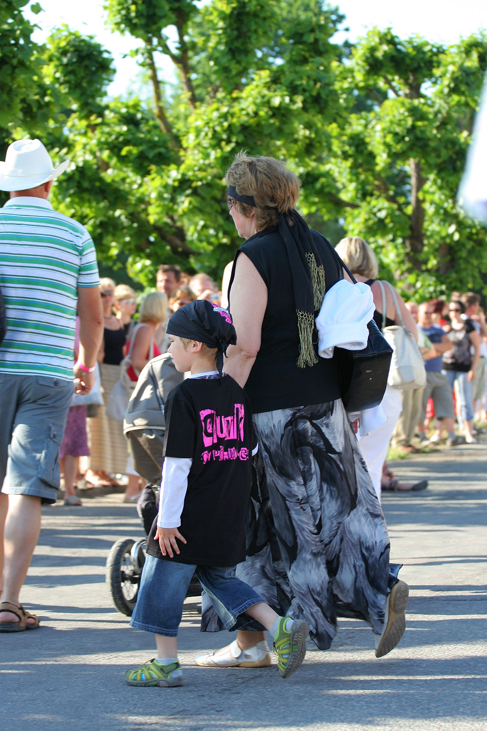 A woman wearing a long, flowing skirt and black top walks hand-in-hand with a child in a black T-shirt and jeans. They’re part of the crowd at Laulupidu, surrounded by greenery and other attendees, some holding bags and pushing strollers. The scene has an eclectic punk vibe woven in with tradition.