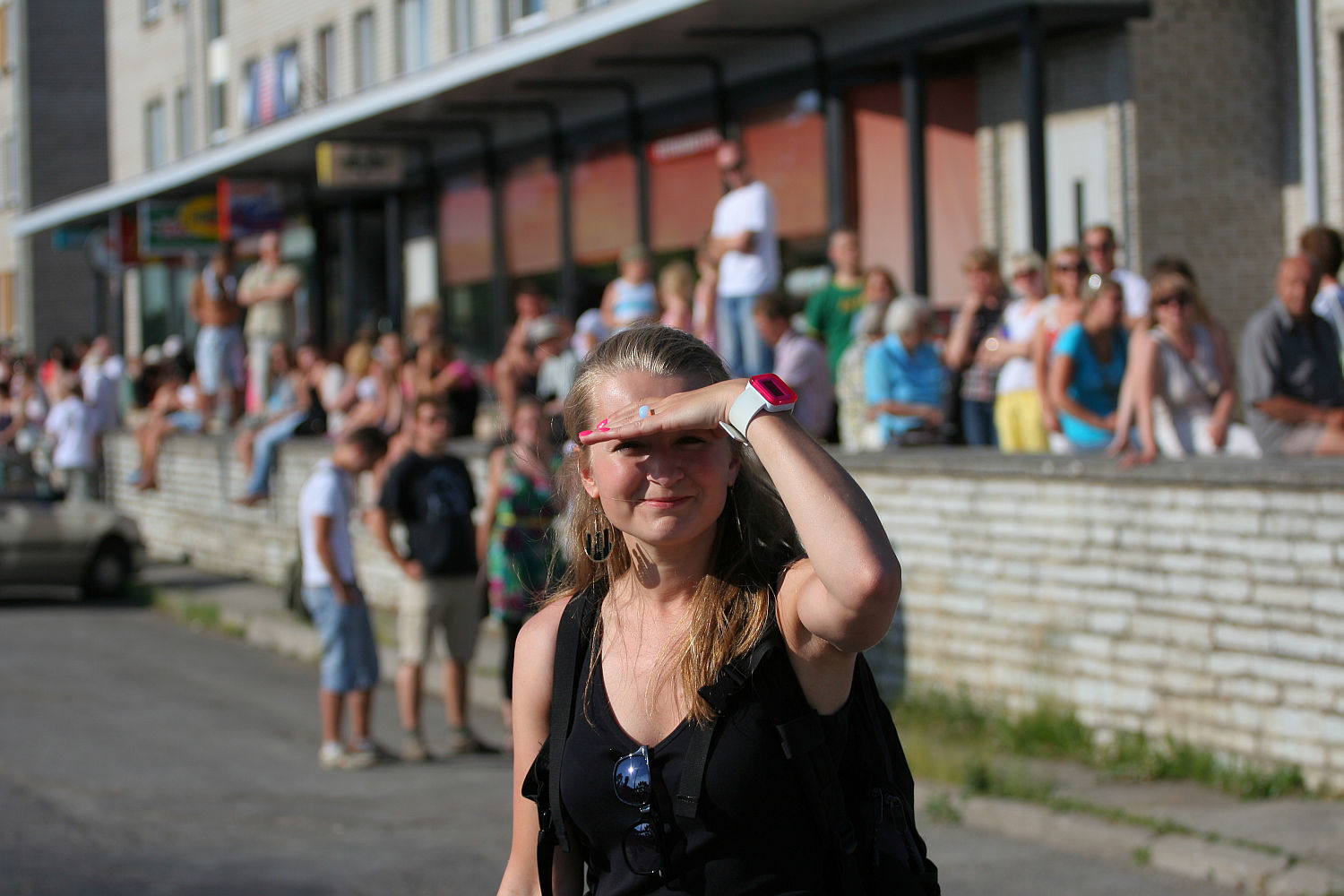 A woman with long hair, wearing a black sleeveless top and sunglasses, is shielding her eyes from the sun with her left hand. She stands on a street in front of a crowd gathered along a brick wall and sidewalk, giving off a punk vibe. The surrounding area includes buildings and parked cars.