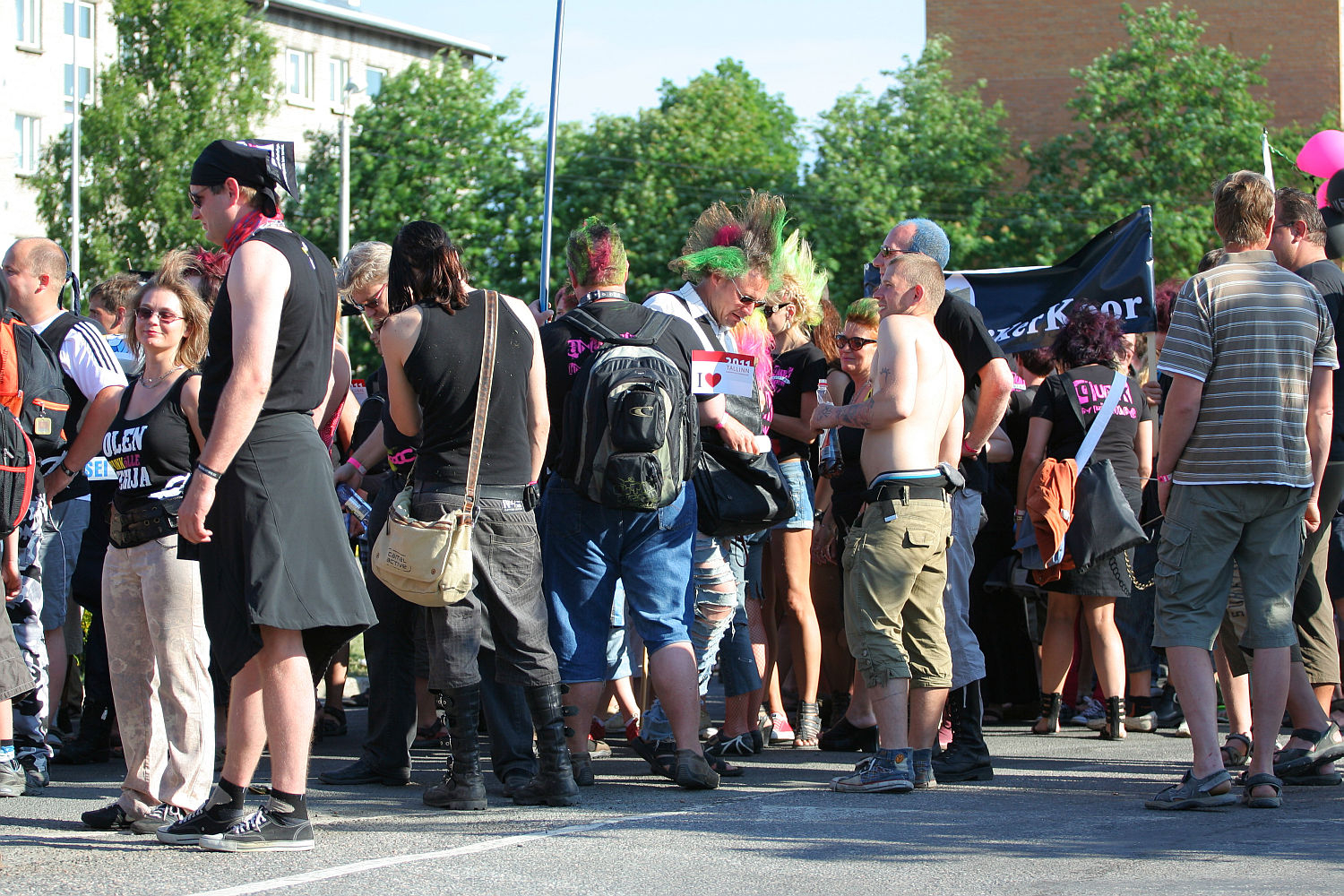 A group of people are gathered outdoors during the daytime at what appears to be a lively Laulupidu. They display various styles, including colorful punk hairstyles, black clothing, and some with bare torsos. The background features trees and a few buildings. A couple of banners are held up among the crowd.