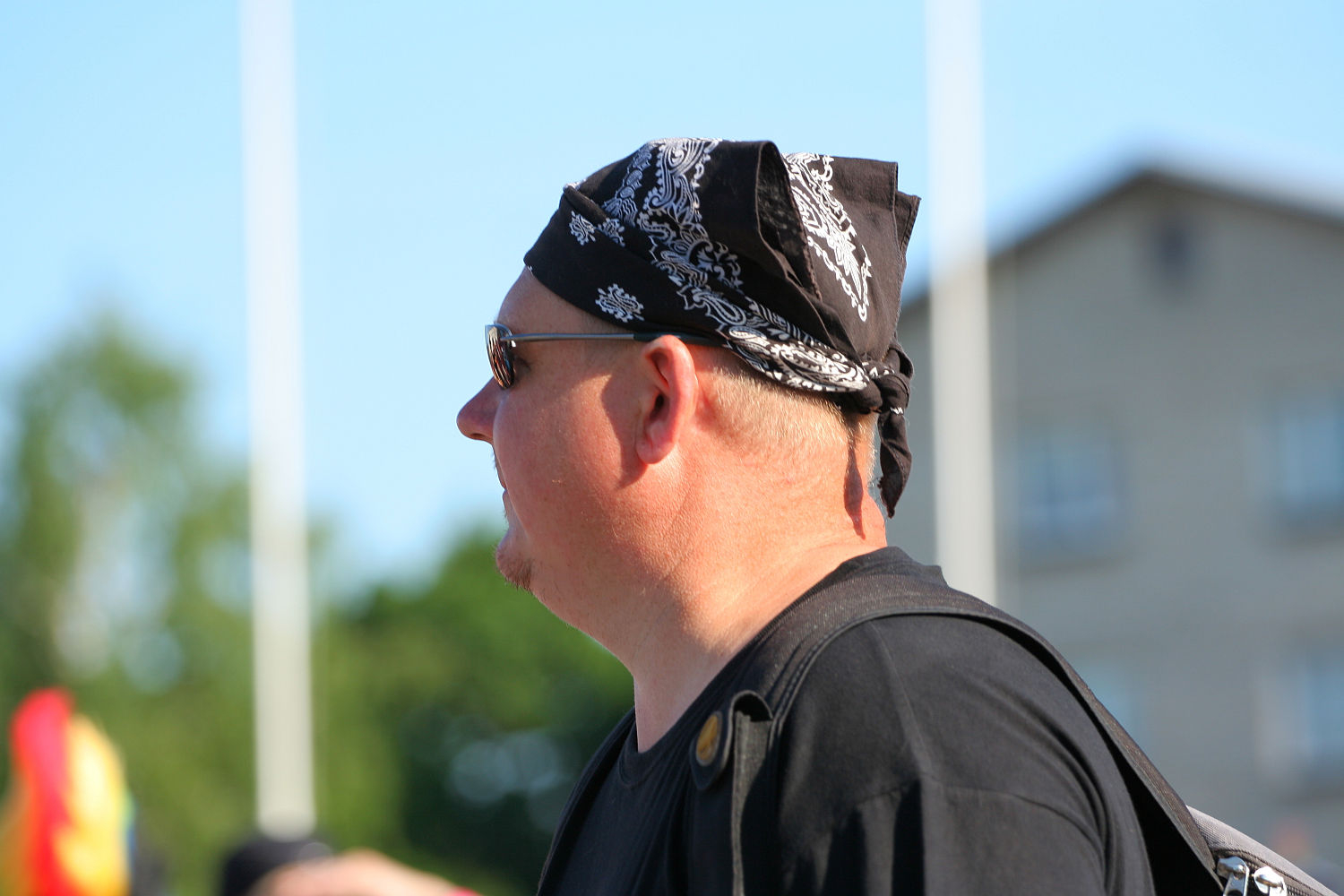 A person wearing sunglasses, a black bandana with a white pattern, and a black shirt stands turned to the side against a backdrop of greenery and a blurred building. With light skin and short hair, they carry a black bag adorned with straps and buttons, exuding an edgy punk vibe reminiscent of Laulupidu.