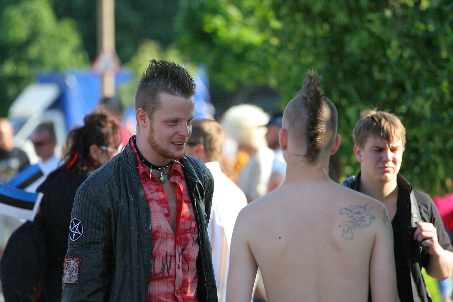 Three men with punk hairstyles and attire are conversing outdoors. The man on the left wears a black jacket with patches and a red shirt, while the man in the middle is shirtless, revealing a back tattoo. A third man stands in the background, wearing a black shirt. The setting resembles Laulupidu festivities in a green park area.