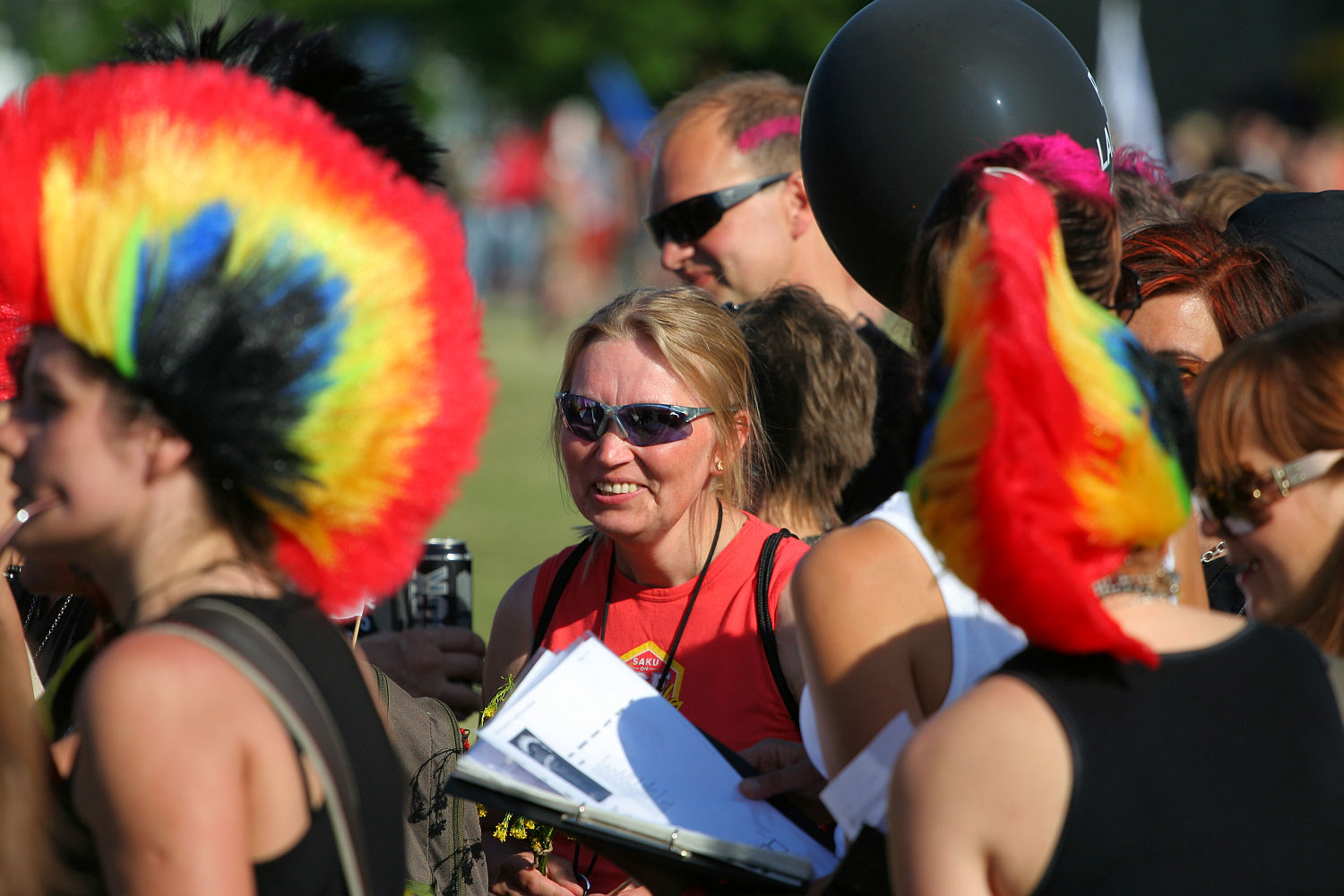 A group of people gather outdoors, many sporting vibrant, punk-inspired rainbow-colored mohawk wigs. A woman at the center, wearing sunglasses and a red shirt, holds papers and a clipboard. The background has trees and a sunny atmosphere, reminiscent of Laulupidu's bright and energetic setting.
