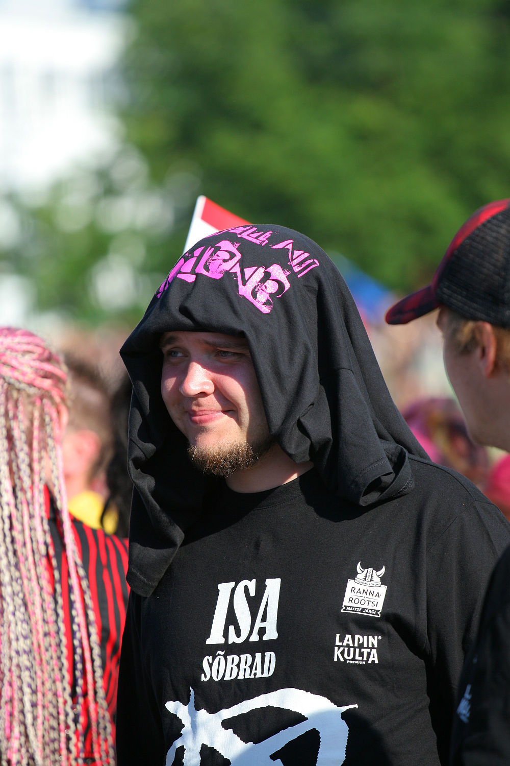 A man with a beard wearing a black shirt with text and logos, and a black shirt draped over his head, stands outside on a sunny Laulupidu day. Other people are visible in the background, with one person having brightly colored punk braids. Trees and a blurred crowd can be seen in the background.