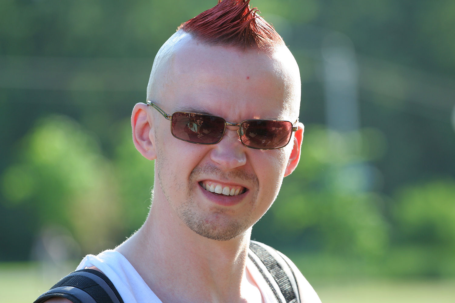 A person with a red mohawk hairstyle, wearing sunglasses, a white tank top, and a black backpack, is seen outdoors on a sunny day. They are smiling broadly, revealing their teeth, with a green, blurred natural background—a true punk spirit enjoying the sunshine.