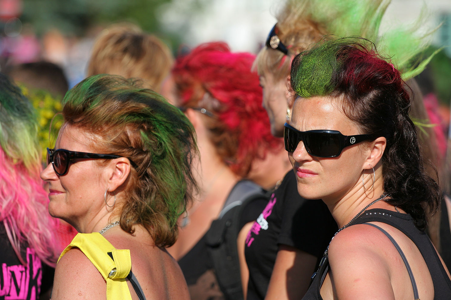 A group of people with colorful, punk-style mohawk hairstyles are standing together outdoors at Laulupidu. Two prominent individuals wear black sunglasses; one has green-tipped hair and the other has green and red hair. The background is filled with more people sporting vibrantly colored, creative hairdos.