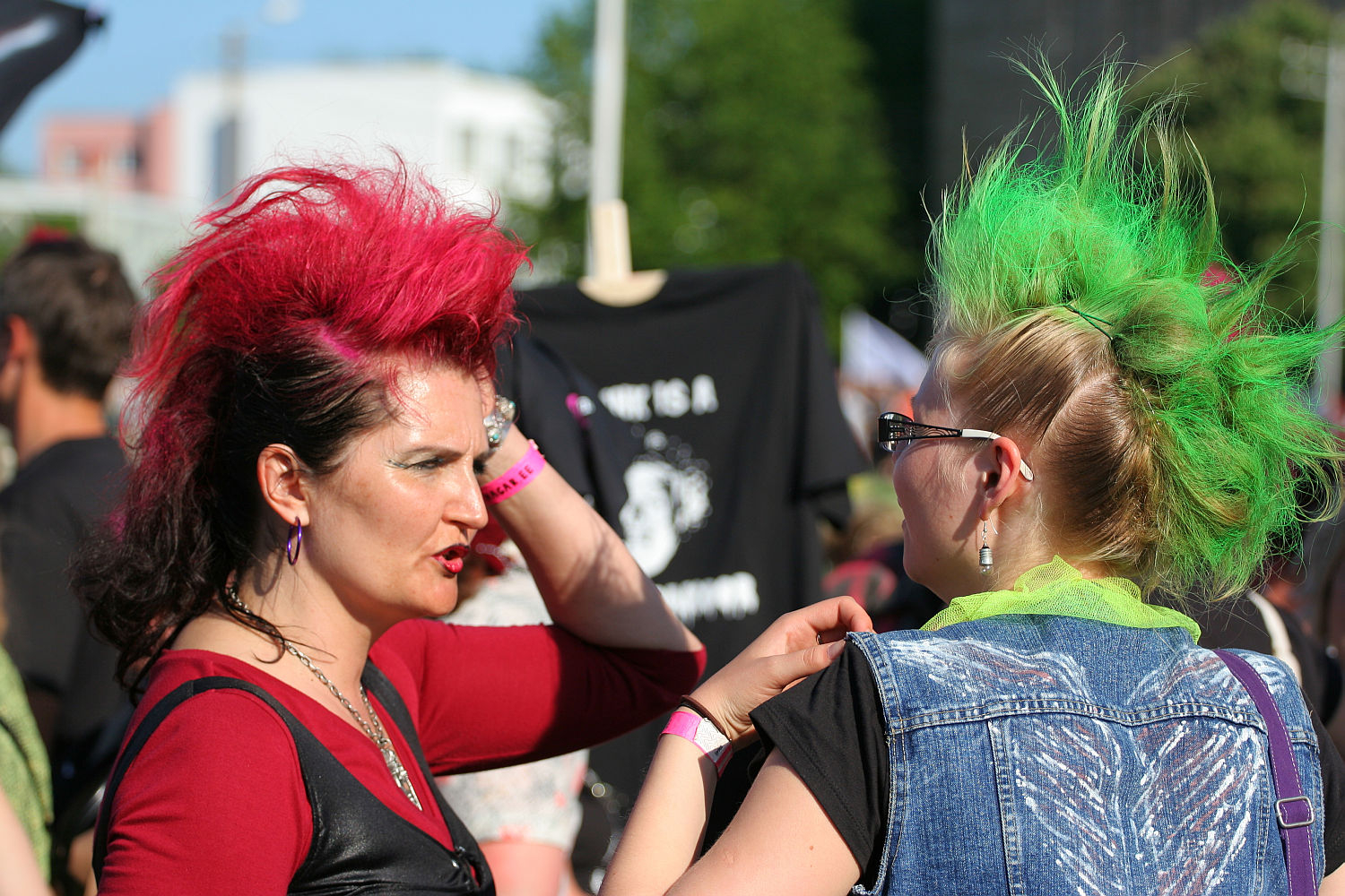 Two women with brightly colored mohawks, one red and one green, are engaged in conversation outdoors. The woman with the red mohawk wears a punk-inspired red top and black vest, while the woman with the green mohawk sports a denim vest and lime green scarf. Both are wearing sunglasses.