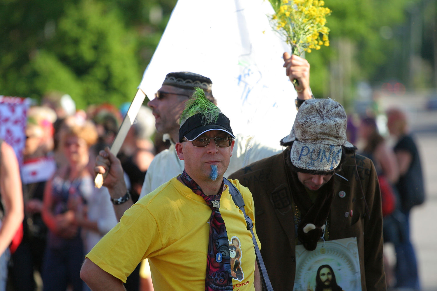 A man with a green mohawk, sunglasses, and a yellow shirt stands in the foreground of a lively Laulupidu procession. Sporting a colorful tie and blue face paint, he exudes Punk vibes. Behind him, other participants carry flags and signs, including one of Jesus. Another man holds yellow flowers and a flagpole. Trees line the background.