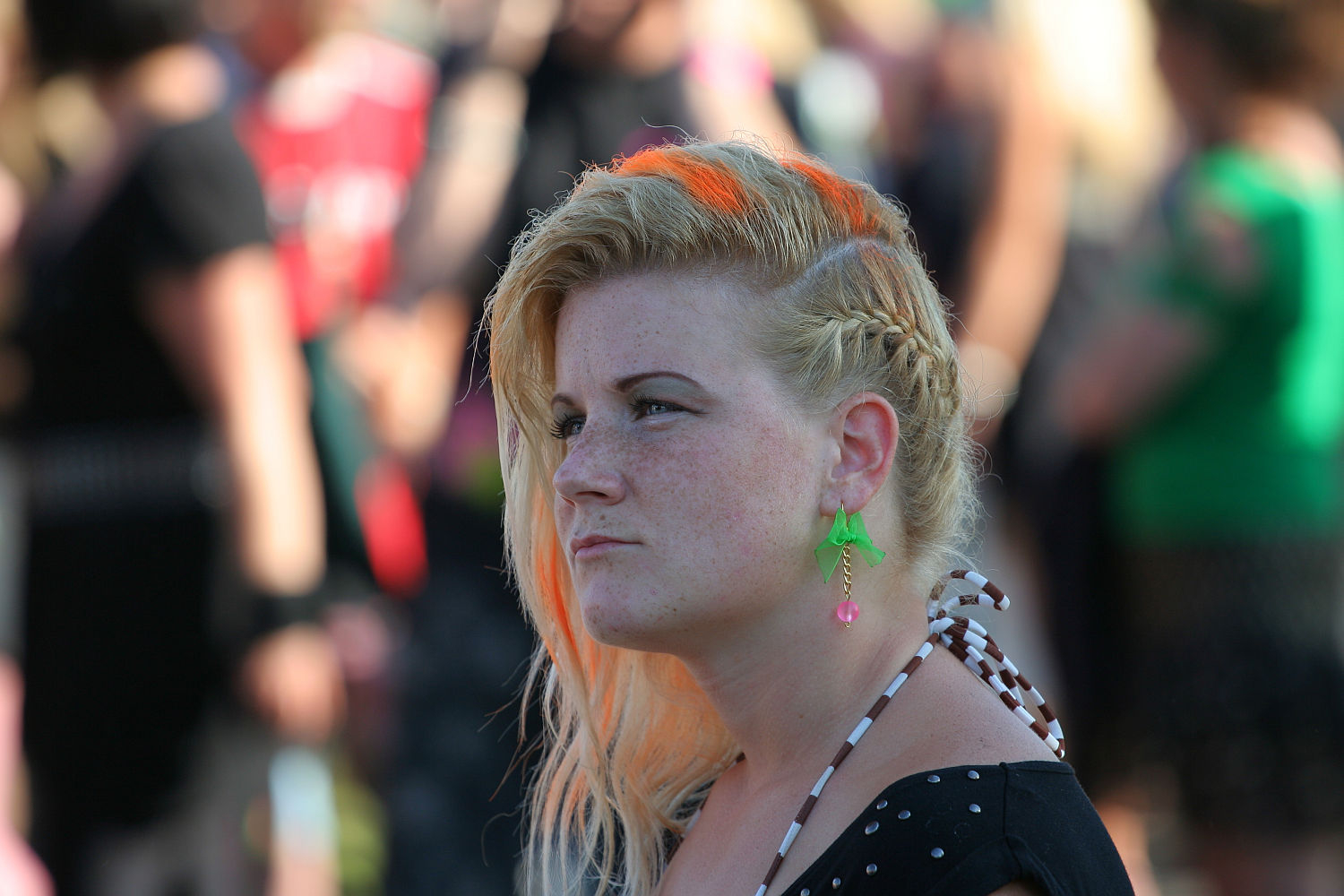 A person with blonde hair featuring orange highlights and a braided side is shown in a close-up. They are wearing green parrot-shaped earrings and have a contemplative expression. The background, possibly at Laulupidu, is blurred with various people and bright colors. The person is wearing a black dotted outfit with straps.