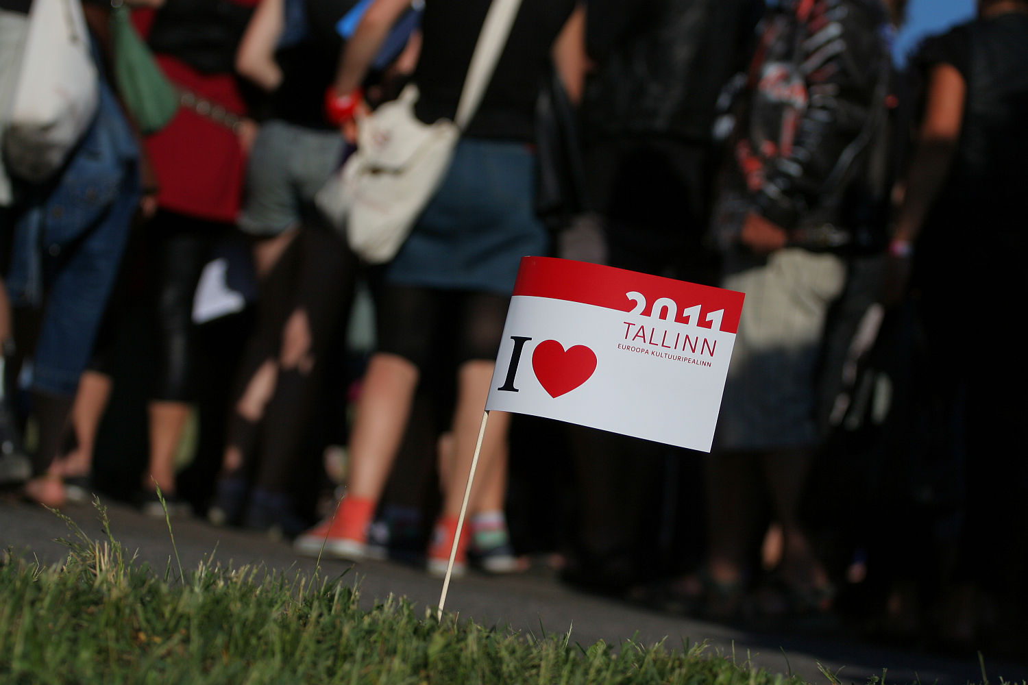 A small flag with the text "2011 Tallinn European Capital of Culture" and "I ♥ Tallinn" is planted in grassy ground. In the background, a group of people, whose lower bodies are visible, stand closely together in casual attire, some wearing backpacks and bags as if ready for Laulupidu.