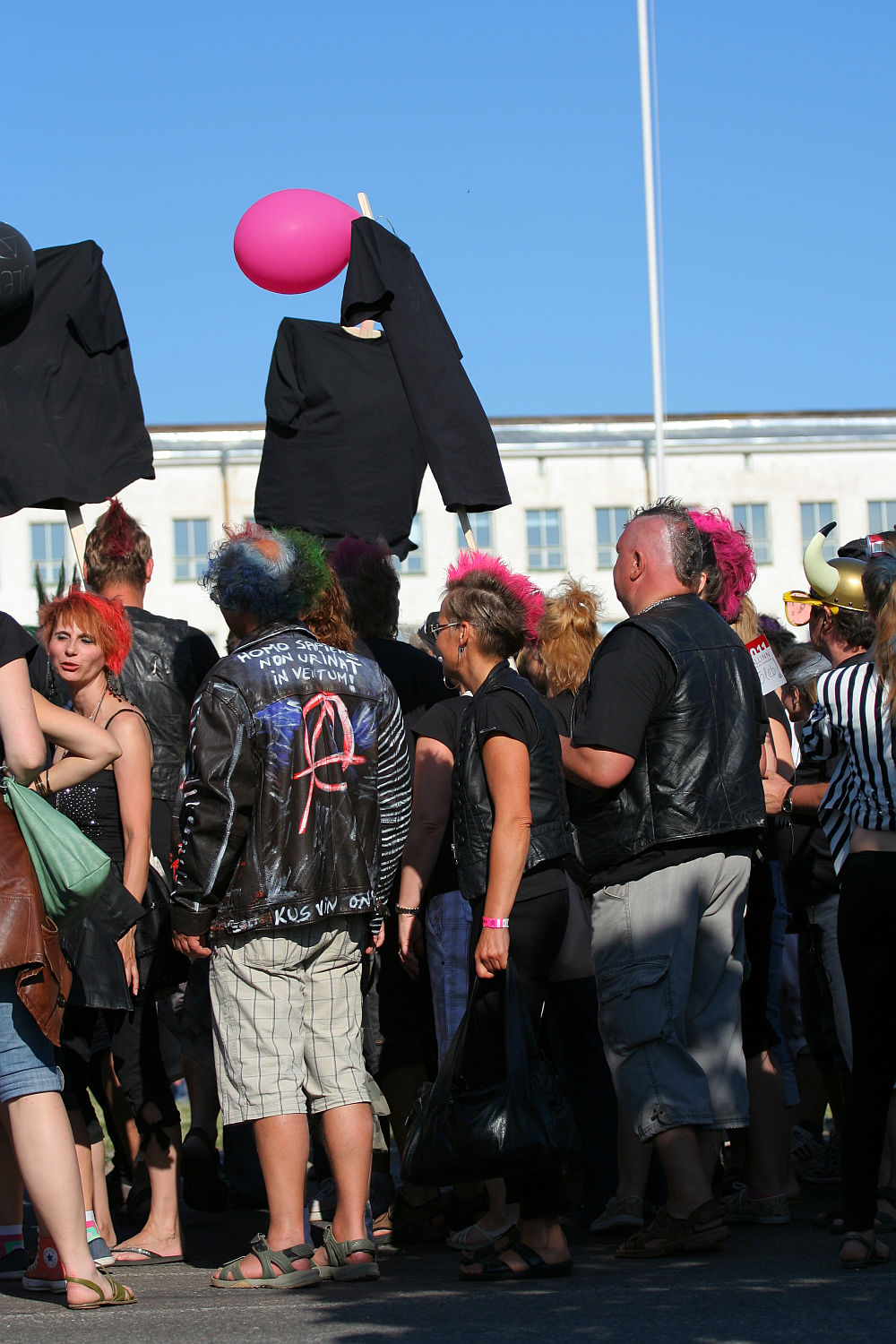 A punk group gathered, many with colorful mohawks and leather jackets, participate in a lively outdoor Laulupidu. Some hold black clothing and pink balloons. The bright blue sky and a large white building are in the background, adding to the festive atmosphere.