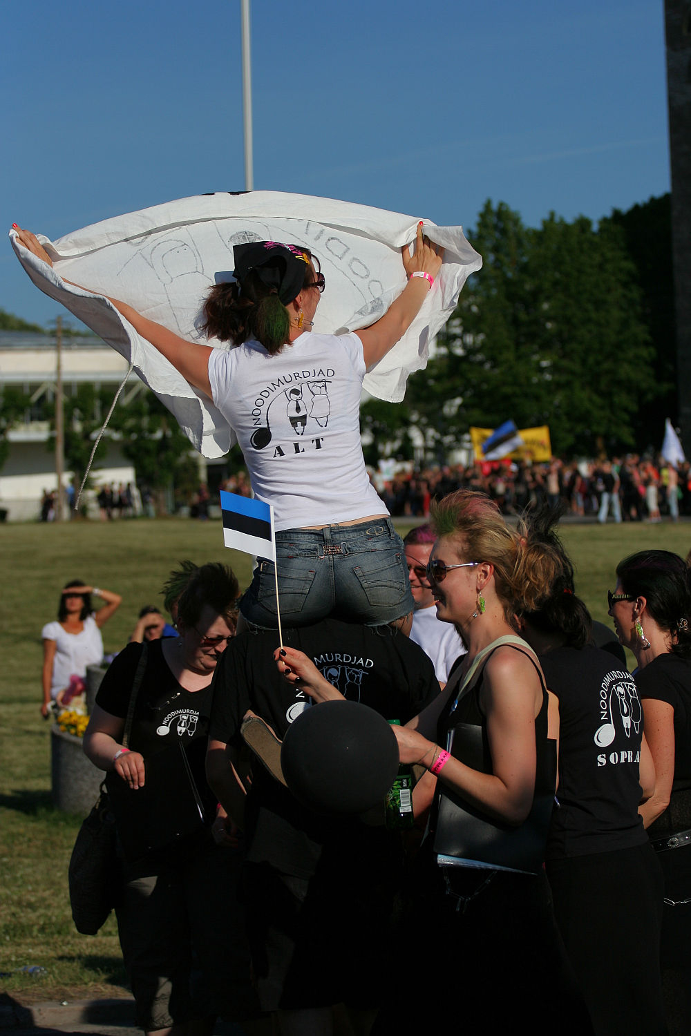 A group of people dressed in black clothing with white logos gathers outdoors on a sunny day, exuding a punk vibe. One woman in the center, wearing sunglasses, stands on another individual's shoulders holding a white cloth above her head. Another person holds an Estonian flag, evoking the spirit of Laulupidu. More people and flags are visible in the background.