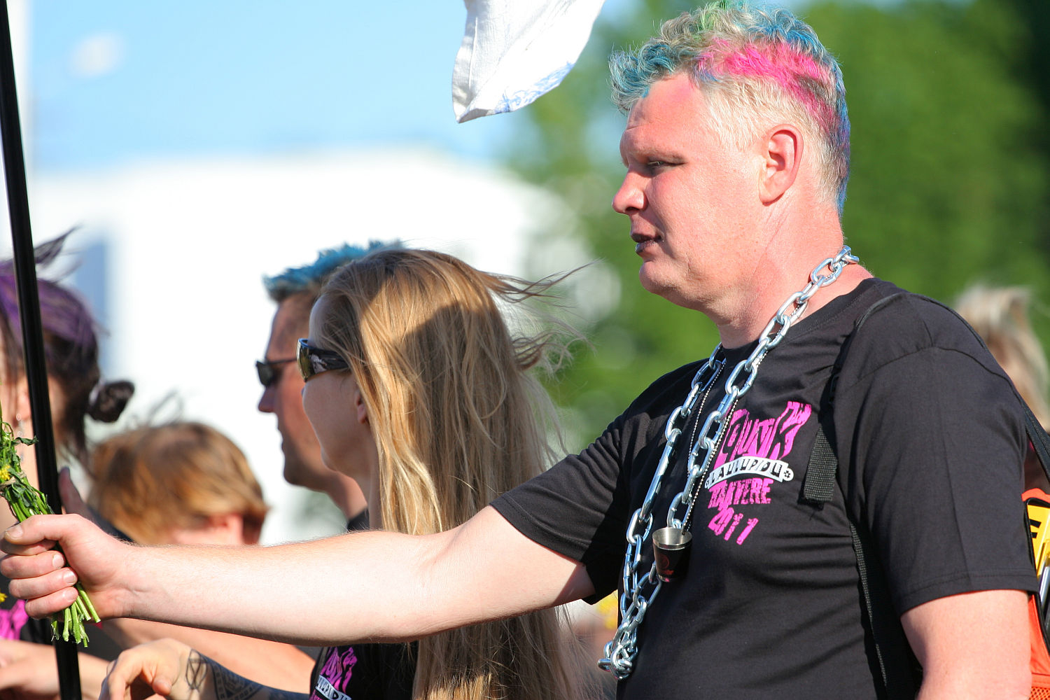 A person with light-colored hair dyed in blue and pink is wearing a black shirt and a large chain necklace, holding a white flag. Sunglasses cover their eyes, giving them a punk vibe. They are part of a group, including a woman with long blonde hair also wearing sunglasses, likely at an outdoor Laulupidu or protest.