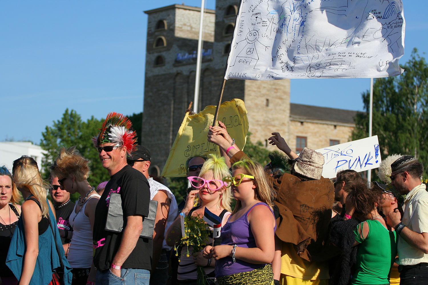 A lively group of festival-goers, some wearing punk-style clothing and accessories, gathers in front of a stone building with two towers. The Laulupidu crowd showcases brightly colored outfits and hairstyles, with some holding signs and flags. The atmosphere is festive and energetic under a clear blue sky.