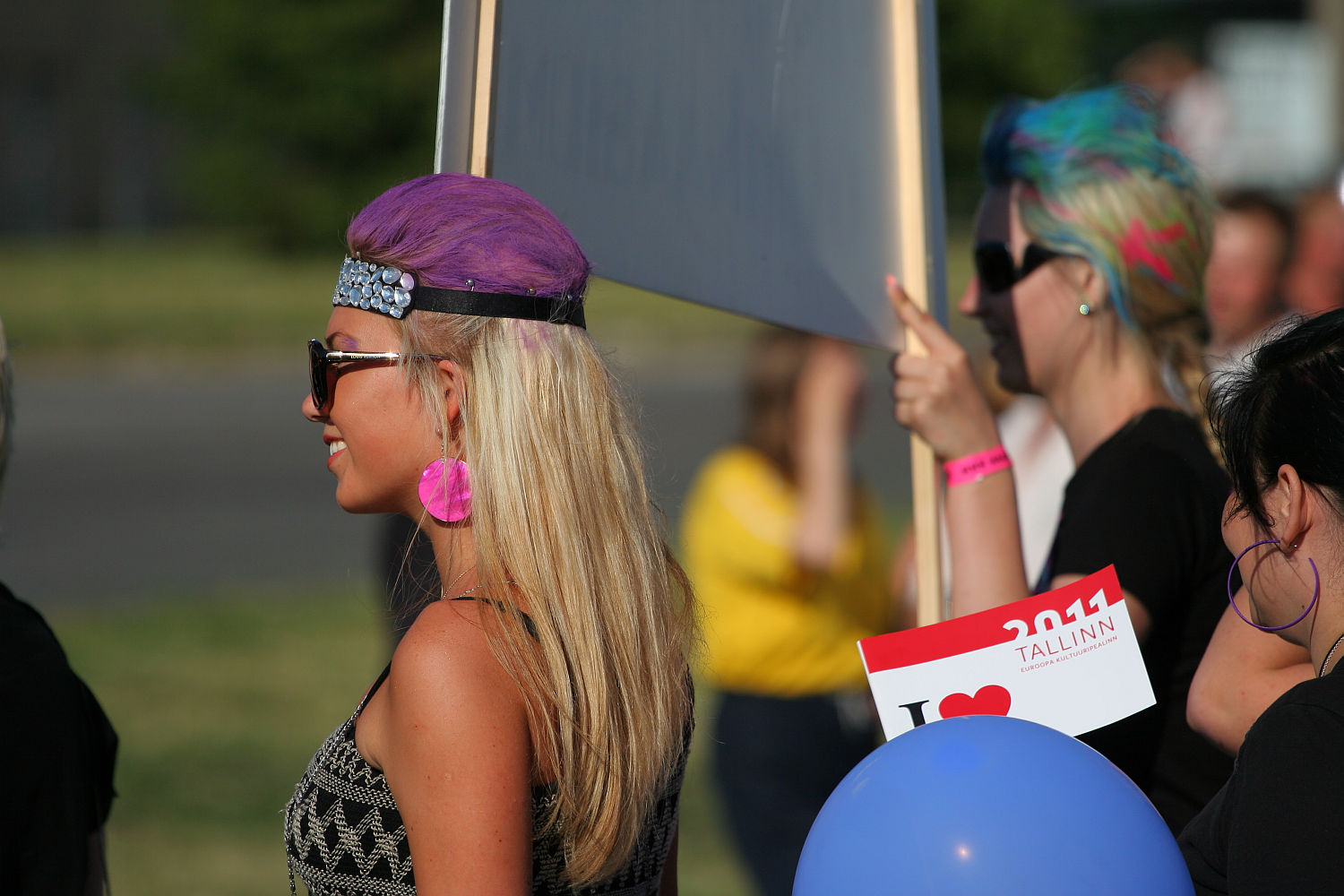 A woman with purple hair and a headband stands outdoors, holding a sign. She wears large pink earrings and sunglasses. Nearby, another punk with colorful hair holds a "Tallinn 2011" sign and a blue balloon. Blurred individuals in the background are in a park-like setting, celebrating Laulupidu.