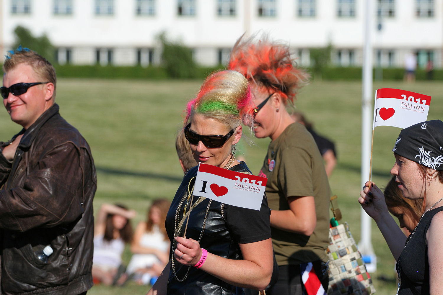 A group of people with colorful punk hair, including neon green and bright orange, stand outdoors holding small flags that read "I ❤️ TALLINN 2011." They're dressed in casual clothing. The background includes a grassy area and a white building. Some seated people are visible in the distance, suggesting a Laulupidu celebration.