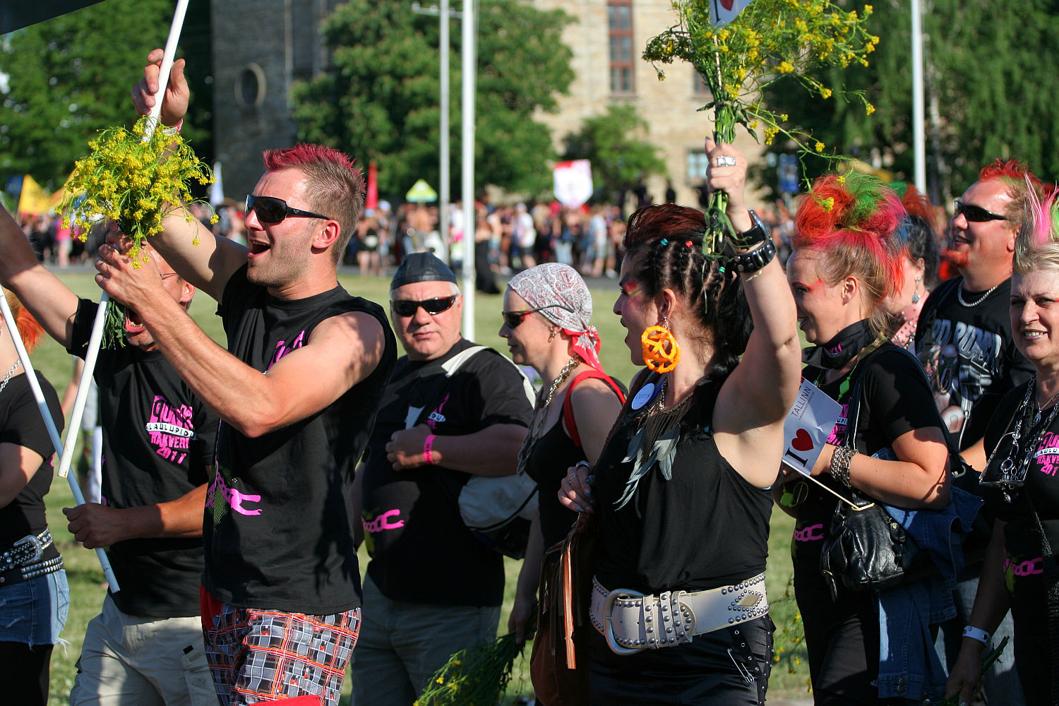 A vibrant group of people, many with dyed hair and colorful clothing, engage in an outdoor celebration or protest. They hold flowers, flags, and signs while smiling and interacting like a punk-themed Laulupidu. The background reveals a crowd and part of a building, indicating a lively public event.