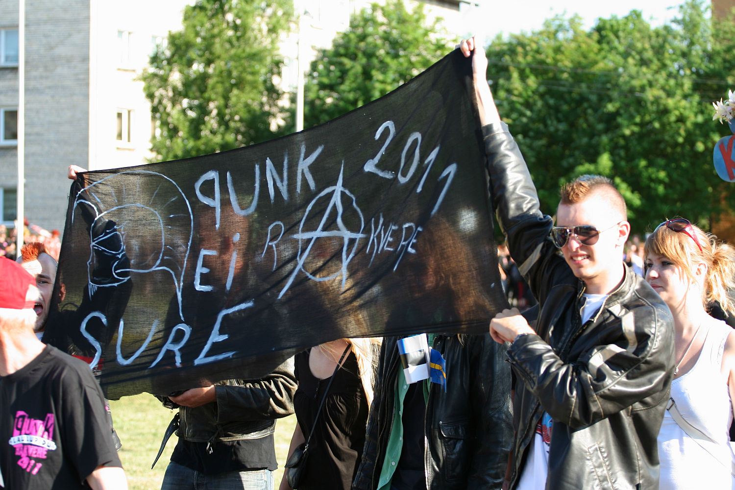 A group of people in punk attire holding a large black banner that reads "PUNK REIR ANKERE 2011 SURE" with various symbols, including an anarchy sign. The individuals, flaunting leather jackets and sunglasses, evoke the spirit of Laulupidu. Trees and a building are visible in the background on a sunny day.