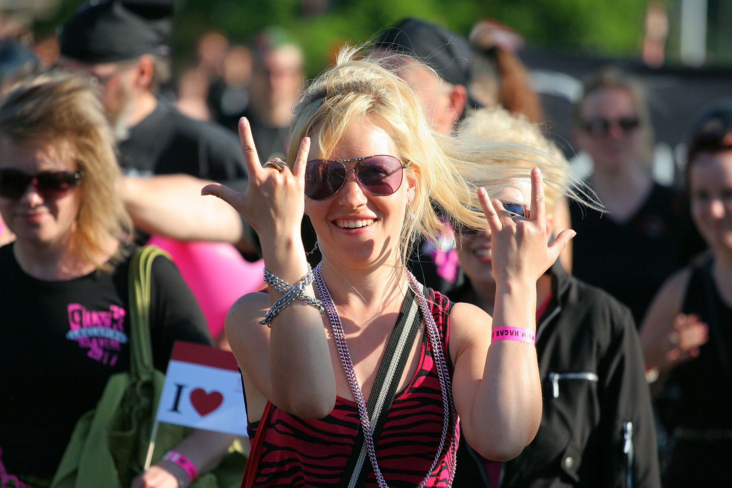 A blonde woman wearing sunglasses gives rock-and-roll hand signs with both hands, capturing the punk spirit. She is smiling and standing in a crowd of people, some of whom are also dressed in black and pink. The crowd appears to be outdoors at Laulupidu, with the woman rocking a striped black and pink top.