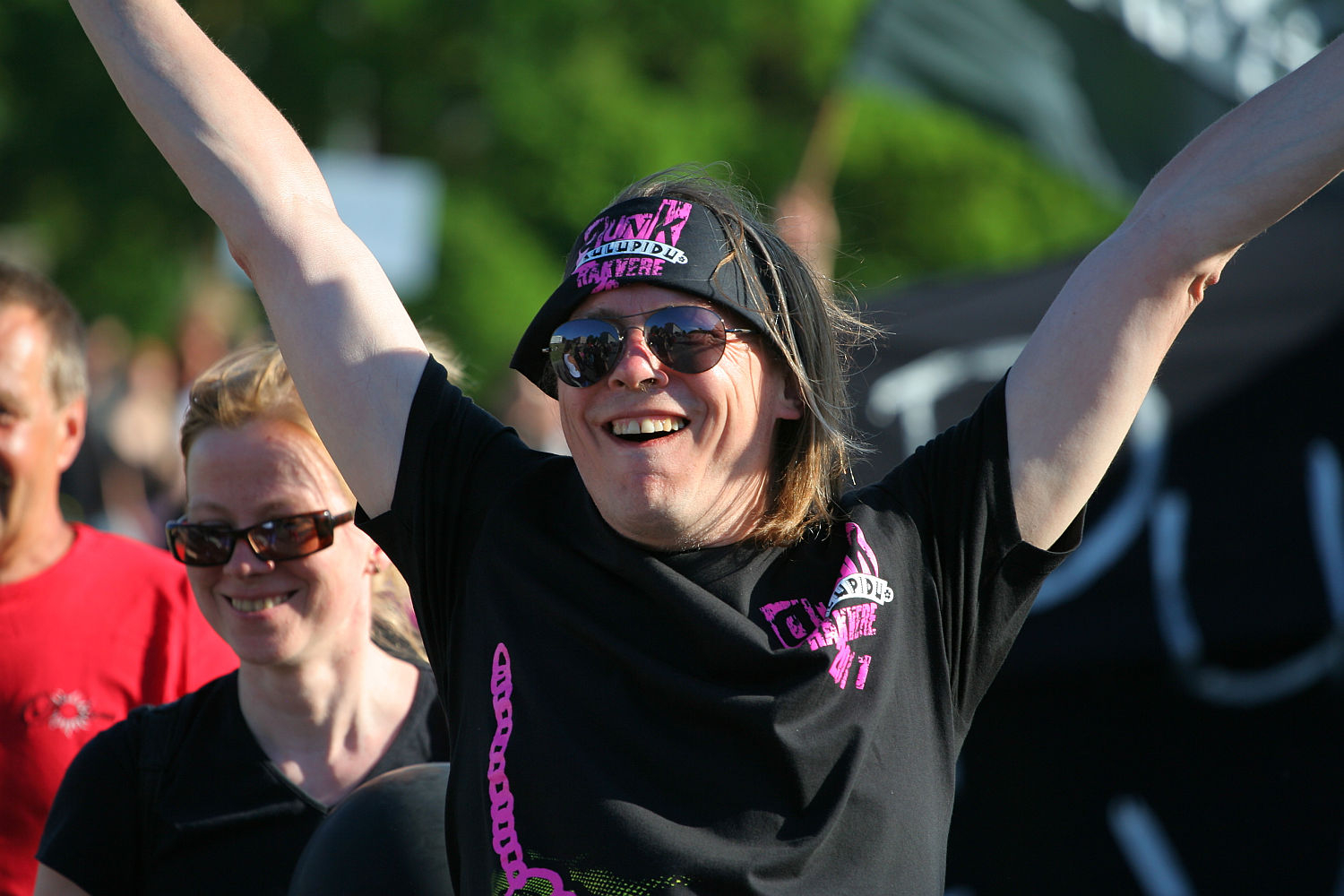 A person with long hair, wearing sunglasses and a bandana, smiles broadly with arms raised in celebration at Laulupidu. They sport a punk-inspired black T-shirt with pink and white graphics. Another smiling individual in sunglasses is partially visible behind them. The background is blurred green foliage and people.