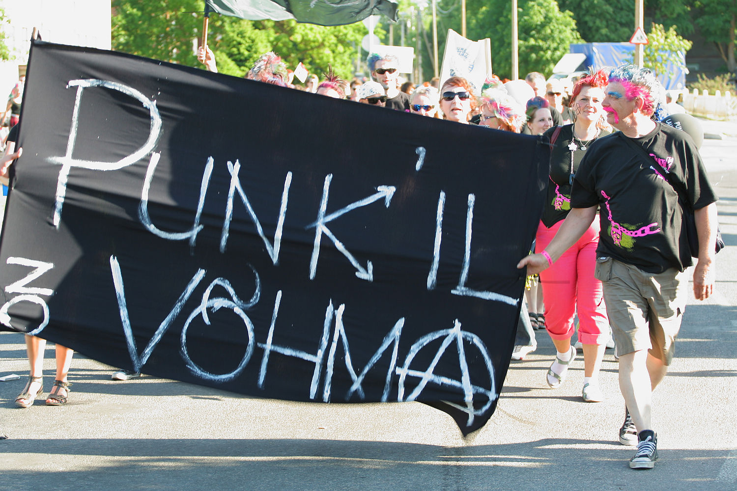 A group of people march outdoors on a sunny day holding a large black banner with white, spray-painted text reading "PUNK IL VÕHMA." The participants, dressed in colorful punk attire with vibrant hair and accessories, embody the spirit of Punk Laulupidu. Trees and other marchers are visible in the background.