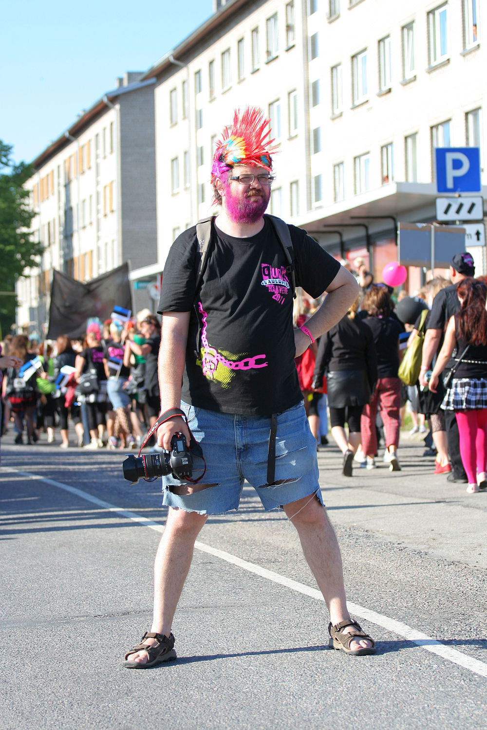A person stands in the middle of the street holding a camera. They sport a colorful mohawk, face paint, and wear a black t-shirt with pink designs, denim shorts, and sandals. A crowd of people and buildings line the background on this sunny day at Laulupidu, capturing the punk spirit perfectly.