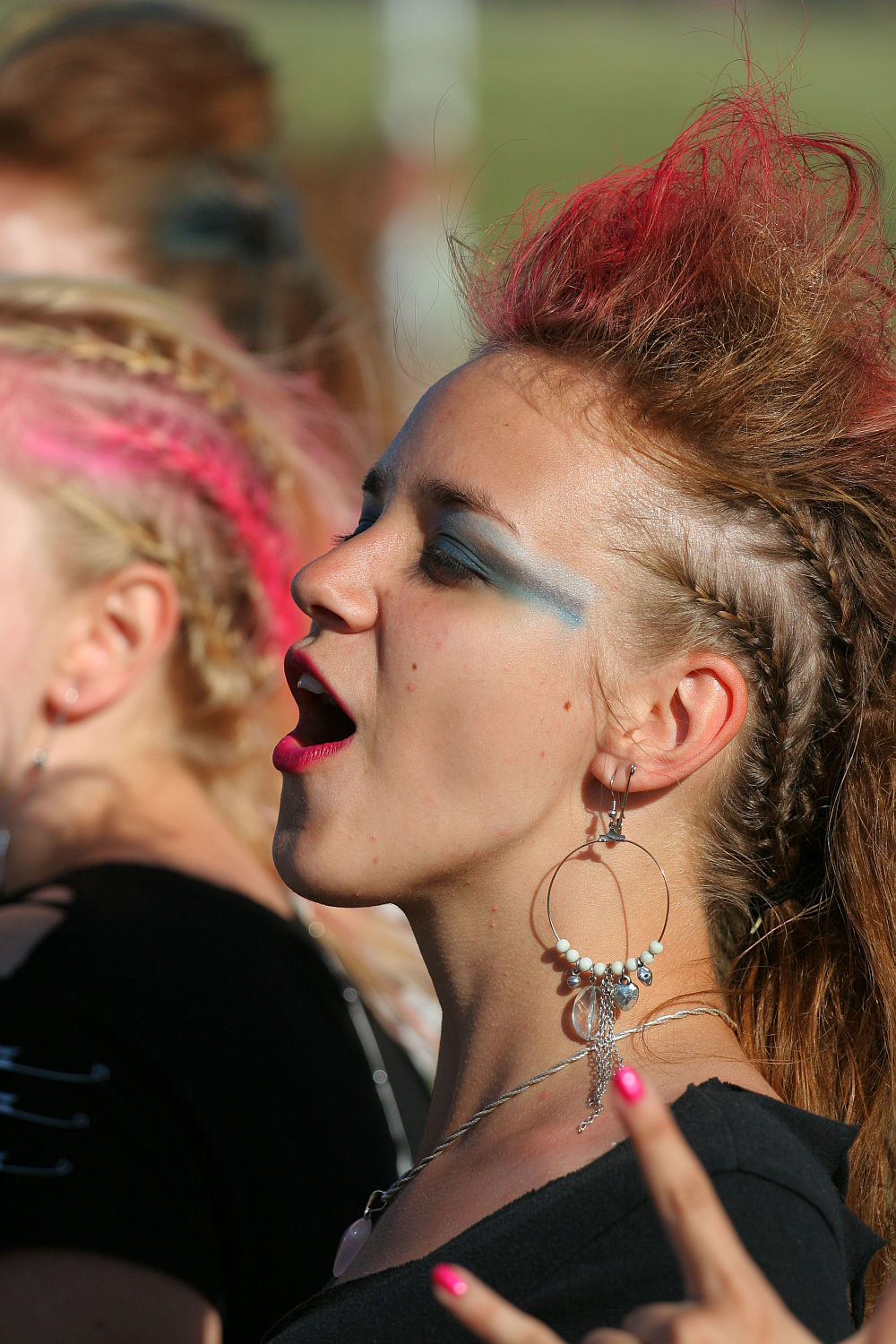 A woman with pink streaks in her hair styled in an updo is singing passionately at Laulupidu. She sports large hoop earrings with multiple dangles and dramatic blue eyeshadow, embodying a Punk vibe. Making a rock on hand gesture, the blurred background suggests an outdoor setting.