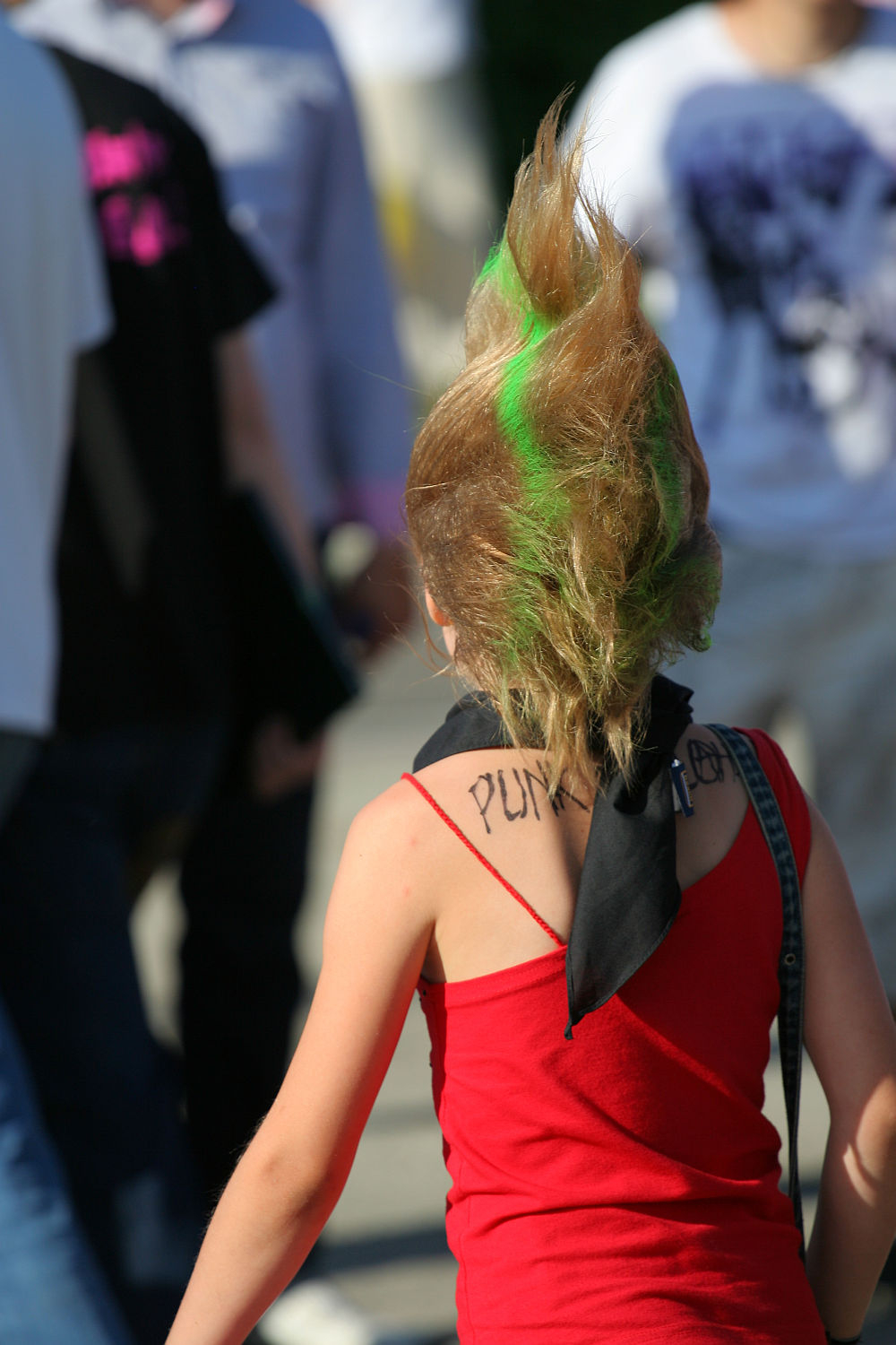 A person with a tall, spiky mohawk dyed with green streaks walks through the crowd at Laulupidu. They are wearing a red tank top and a black bandana with "PUNK" written on it around their neck. The background shows several people dressed casually in various outfits.