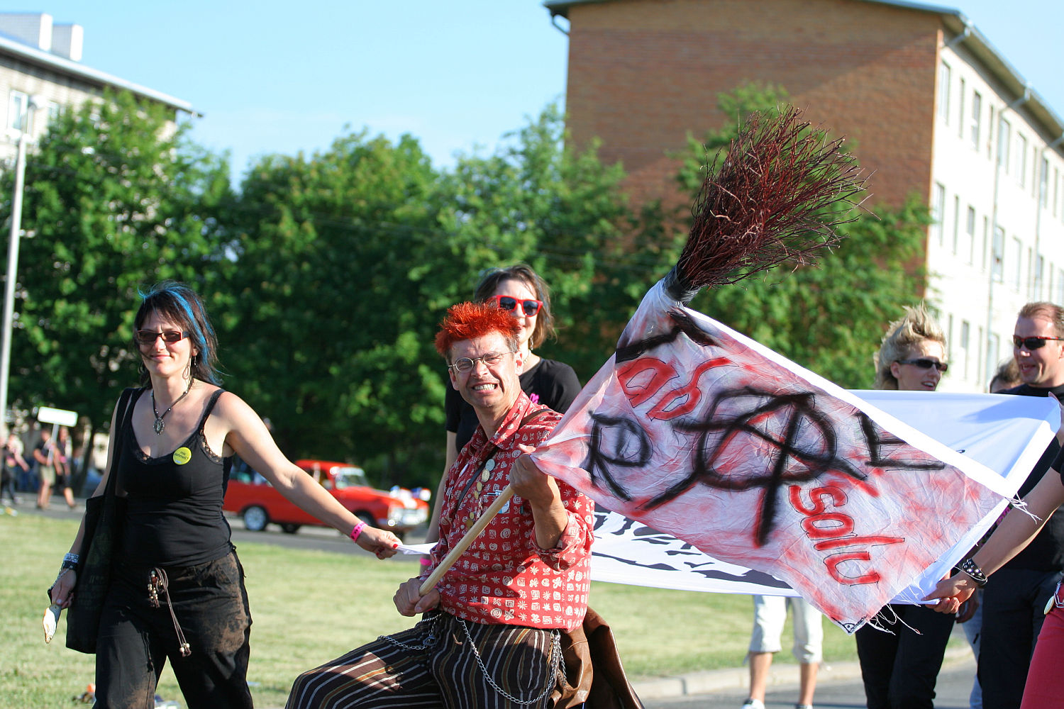 A group of people march outdoors on a sunny day. One person with vibrant red hair and punk-patterned clothing is in focus, holding a flag with anarchist symbols and the words "solidarność". Other participants, dressed in casual attire, walk alongside. Greenery and buildings are visible in the background.