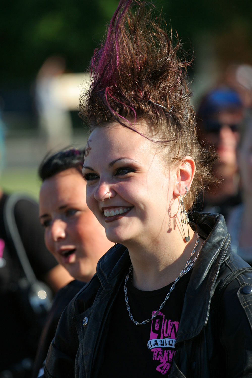 A smiling person with a mohawk dyed purple and black, epitomizing punk style, is wearing a black leather jacket and silver necklace. They have multiple earrings and visible makeup. Another person with a short hairstyle and wearing black can be seen in the background, slightly out of focus, giving the scene a Laulupidu vibe.