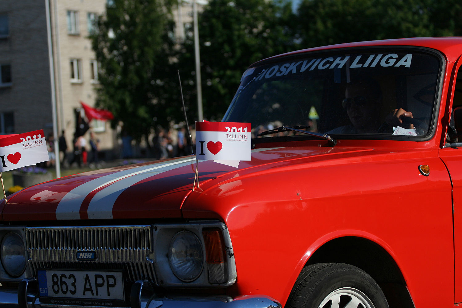 A red vintage car with white racing stripes on the hood is parked. It has several small flags with "I ❤️ TALLINN" and "2011" printed on them, giving it a punk vibe. The front license plate reads "863 APP." The car, part of a gathering akin to Laulupidu, is seen with other cars and people in the background.