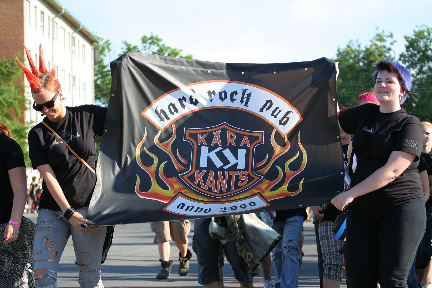 A group of people walking outdoors and holding a banner for "Kära Kants," a hard rock pub established in 2000. The punk-themed banner features flames around a shield-like logo. Participants wear black clothing; one person has a prominent red mohawk, and another has colored, spiked hair. Trees and buildings are visible in the background.