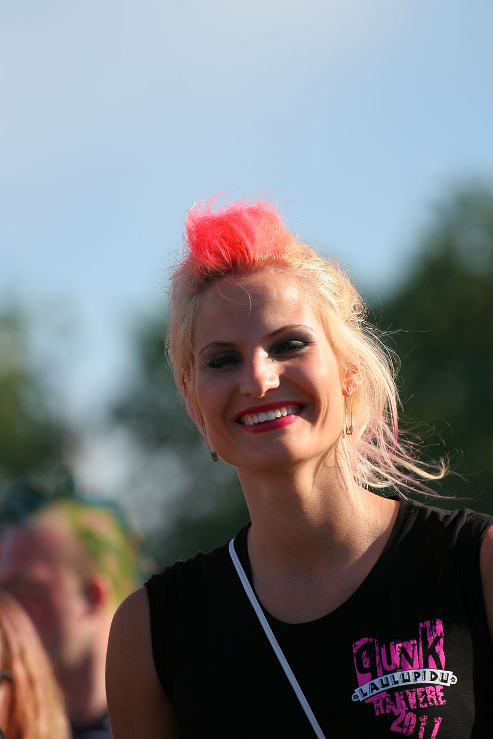 A woman with short, platinum blonde hair featuring a bright pink section at the top is smiling. She is wearing a sleeveless black top with a colorful design on it. The background is an outdoor setting, possibly an Estonian music event, with a clear sky and trees blurred in the distance.