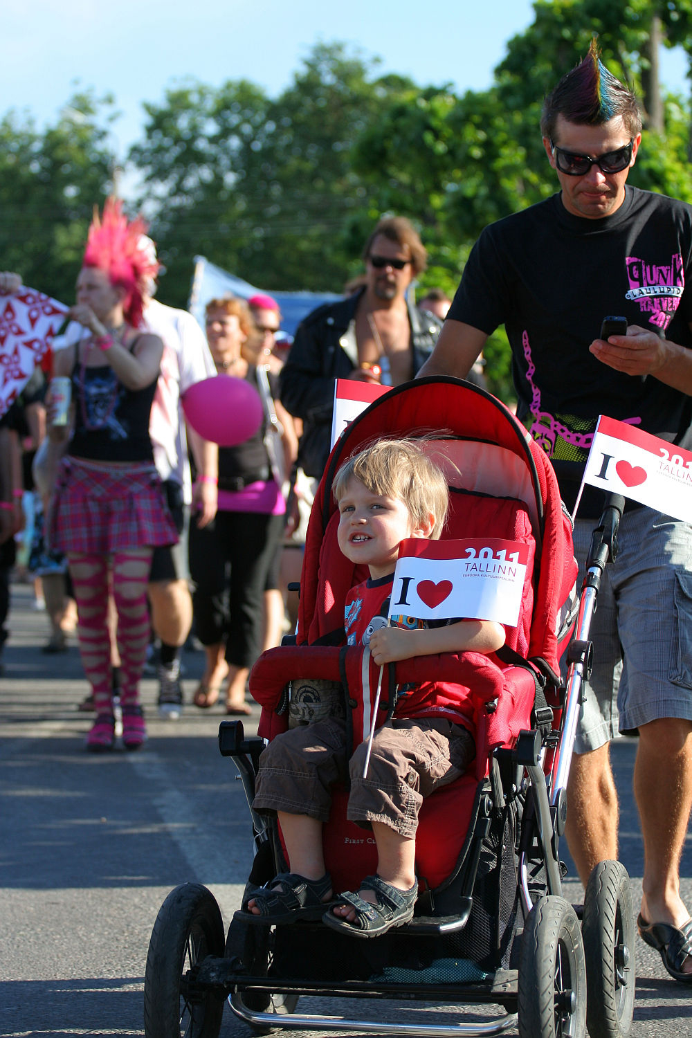 A young child in a red stroller holds a small "I ♥ Tallinn" flag in each hand. The child is pushed by a man wearing sunglasses and a black t-shirt. They are part of the lively Punk Laulupidu street parade, with participants in colorful and eclectic outfits, including pink plaid skirts and flashy accessories.
