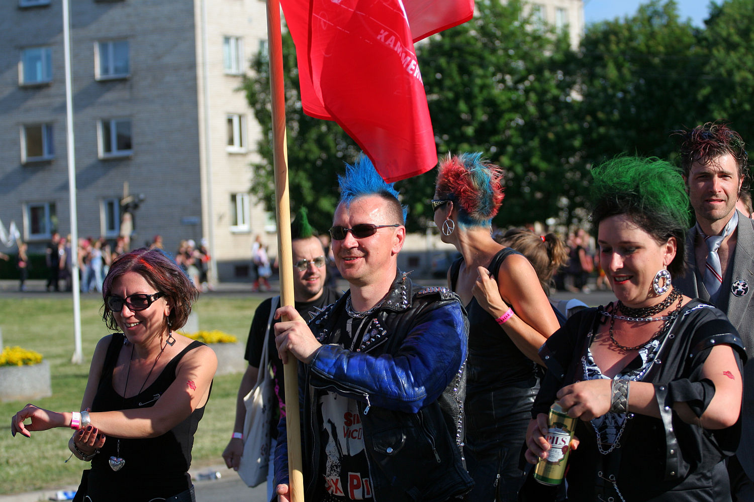 A group of people with colorful mohawk hairstyles and punk outfits are walking outdoors, holding a red flag. They seem to be part of a Punk Laulupidu, with onlookers and buildings visible in the background. Some are smiling and appear to be in high spirits.