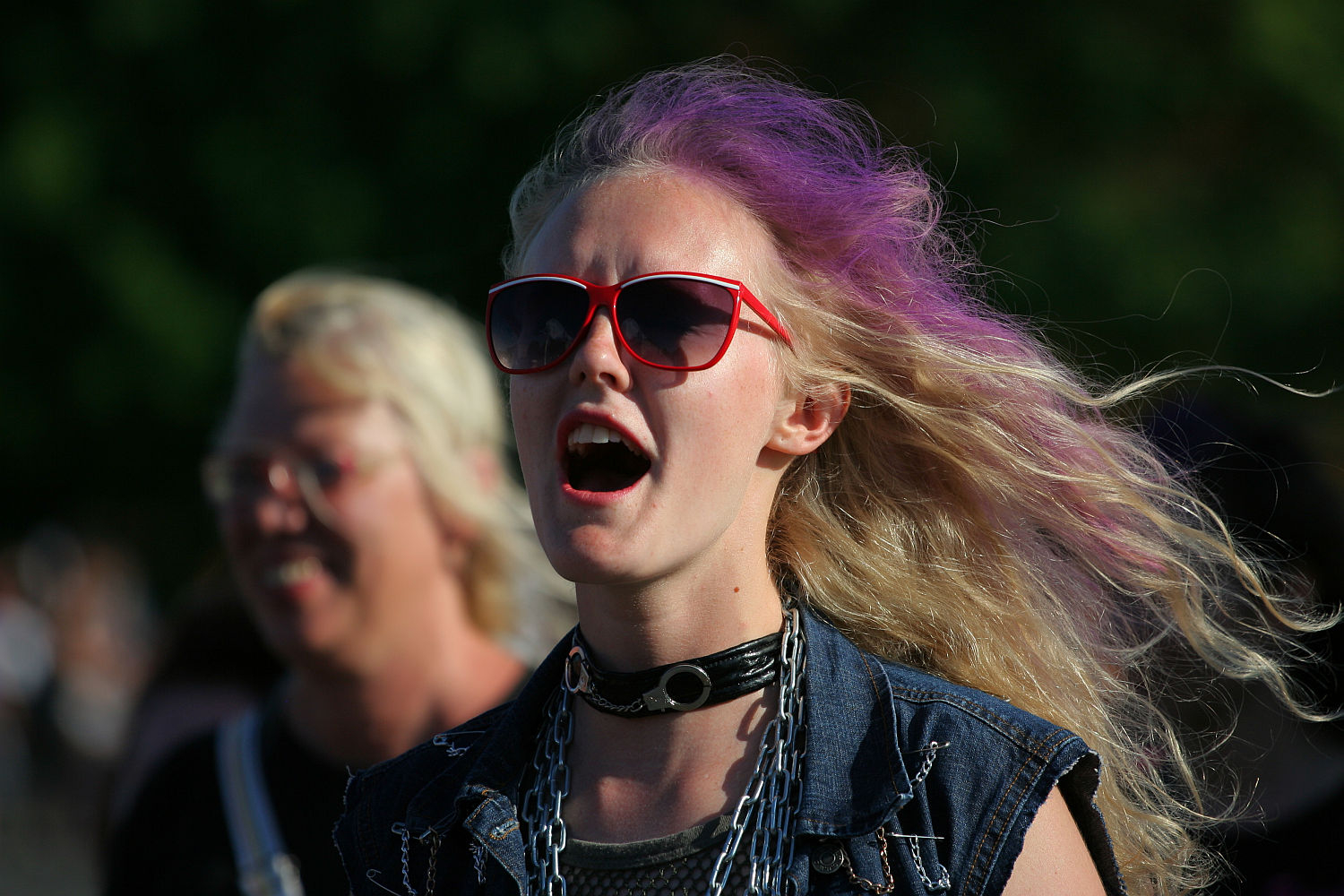 A person with long, wavy, blonde hair partially dyed purple is wearing red-framed sunglasses and a black choker with a circular ring. They appear to be mid-speech or singing with their mouth open, evoking the spirit of Laulupidu. Dressed in a denim vest, they stand outdoors with another individual blurred in the background.