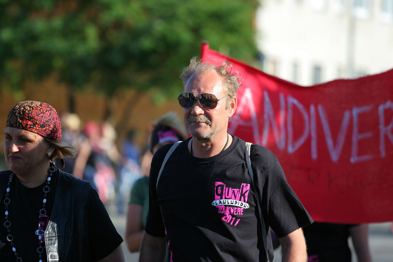 A group of people walk in a street parade. The man in the center wears sunglasses, a black t-shirt with colorful text, and carries a backpack. A woman next to him, with a punk vibe, wears a bandana and necklace. In the background, another person holds a red banner that partially reads "DIVER." Trees are in the distance.