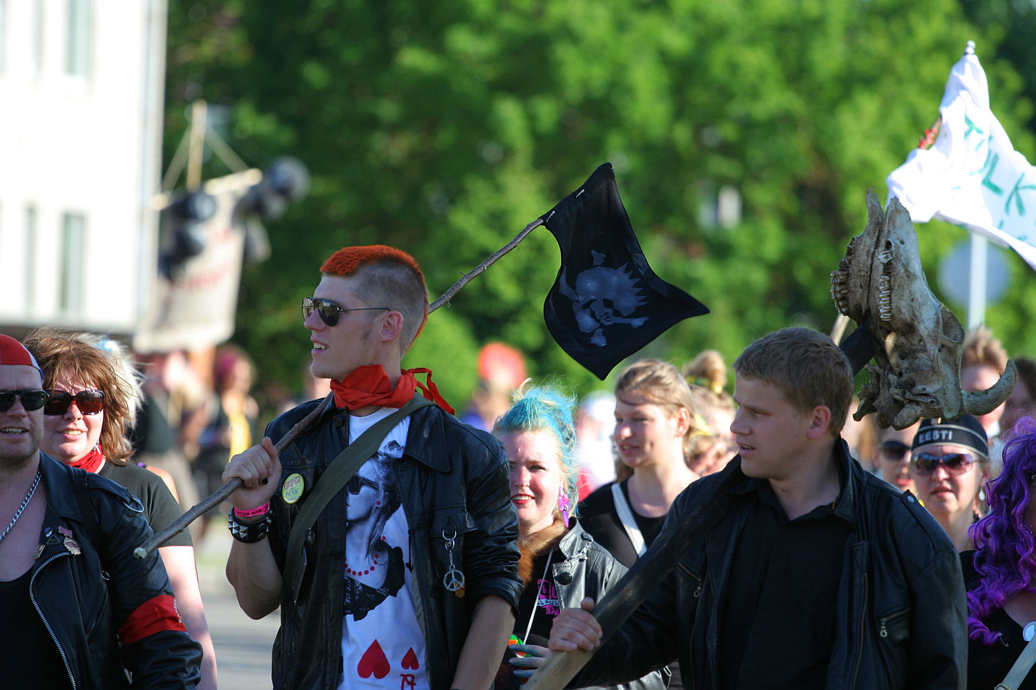 A group of people, some with brightly colored hair and black leather clothing, are marching outdoors. One person with short red hair carries a black flag with a skull design, evoking a punk Laulupidu vibe. Another person carries an animal skull on a stick. Trees and a building are visible in the background.