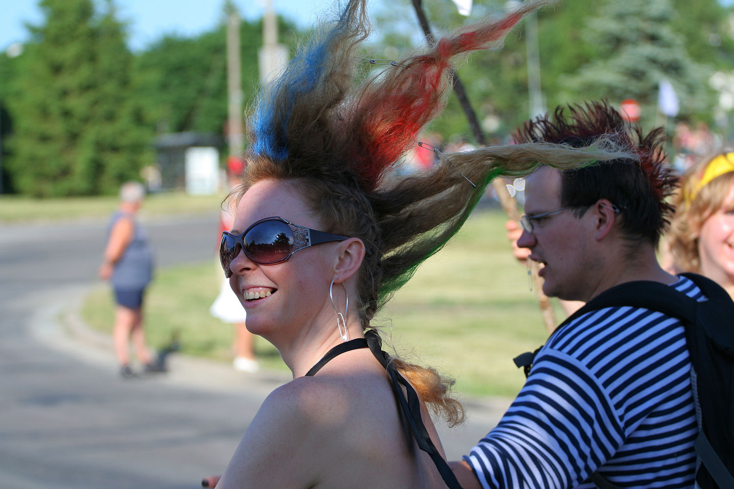 A punk with colorful dyed hair and sunglasses smiles while standing outdoors. Their hair is styled in spikes with red, blue, and green colors. They wear large earrings and a halter top. Another person in a black and white striped shirt stands next to them. Trees and other people are visible in the background, giving a festive Laulupidu vibe.