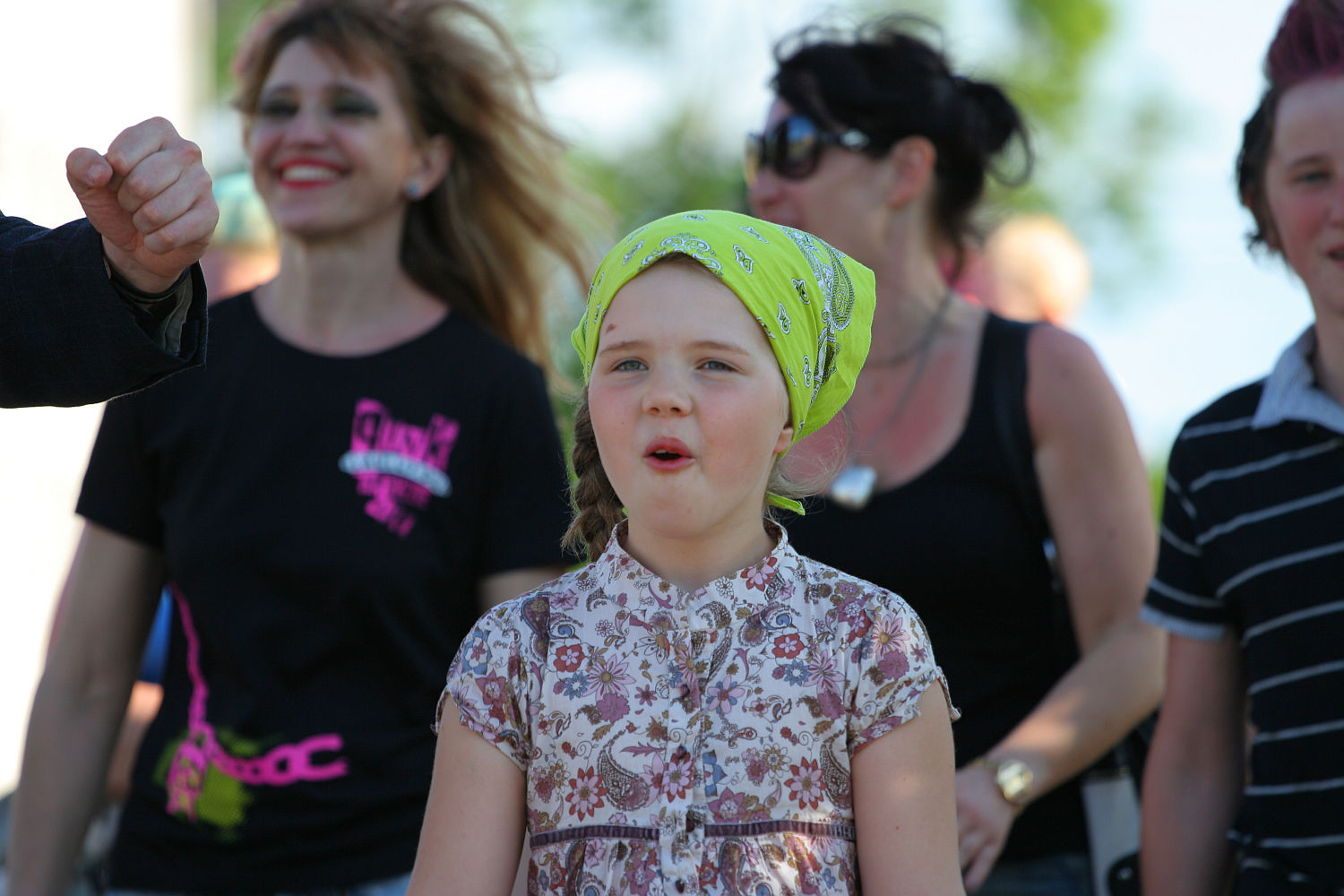 A young girl wearing a floral dress and a yellow bandana stands outdoors with her mouth slightly open, bringing an unexpected punk flair to the scene. Behind her, several adults and another child are visible, some smiling. The background is slightly blurred, suggesting movement and a lively Laulupidu atmosphere under bright sunlight.