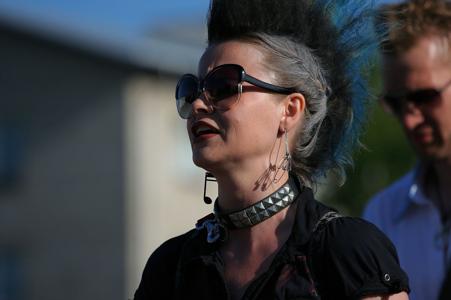 A person with a tall black and grey mohawk hairstyle is seen outdoors, embodying a punk aesthetic. They wear large, dark sunglasses, long earrings, and a studded choker necklace. The background shows blurred buildings and greenery, with another person partially visible behind them. The lighting suggests it is daytime.