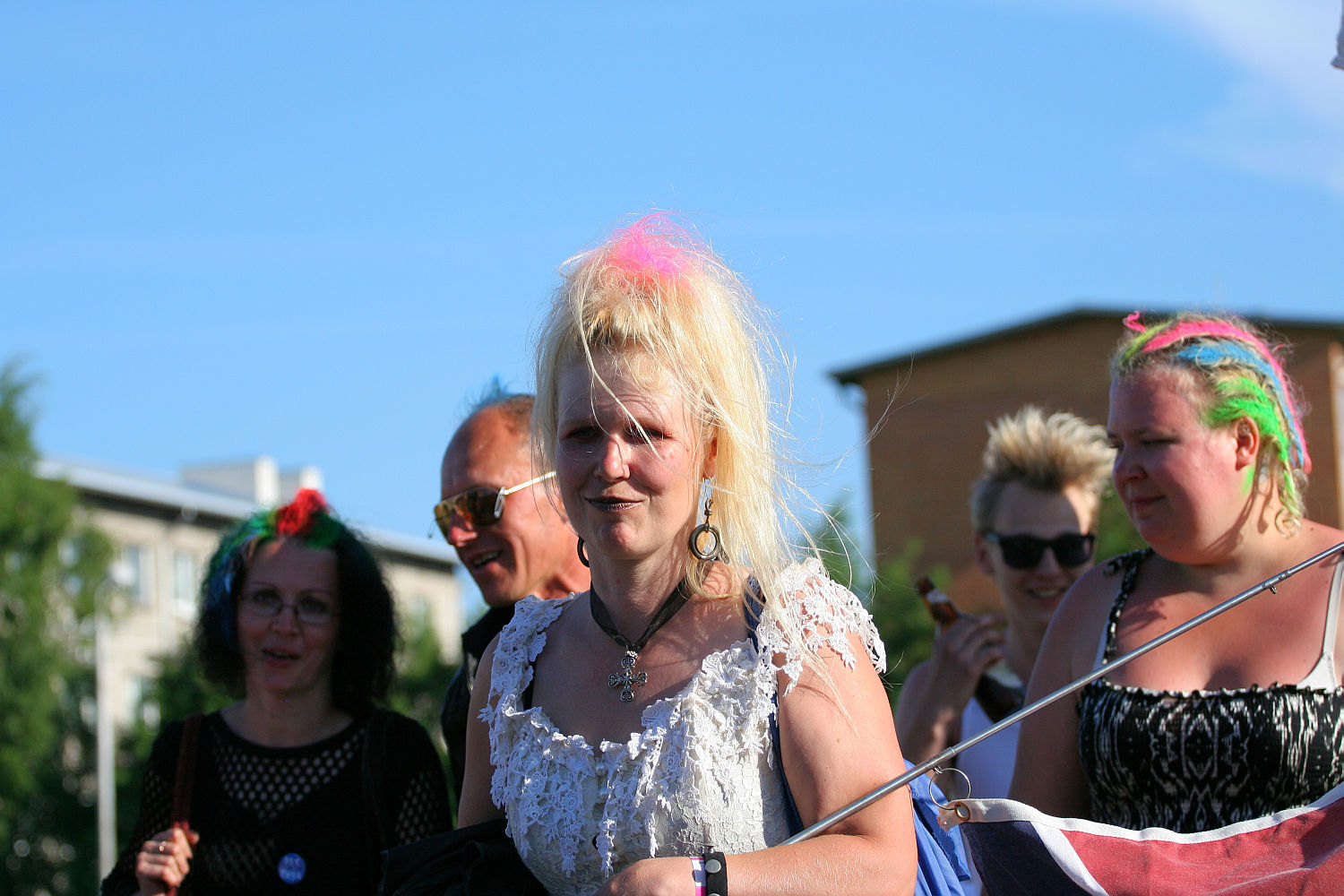 A group of people with colorful, punk-inspired hair, some sporting spiked and dyed styles, are enjoying an outdoor event on a sunny day. A woman in the foreground wears a white lace top, with pink and blue streaks in her hair. The lively scene resembles the festive spirit of Laulupidu against a backdrop of buildings and blue sky.
