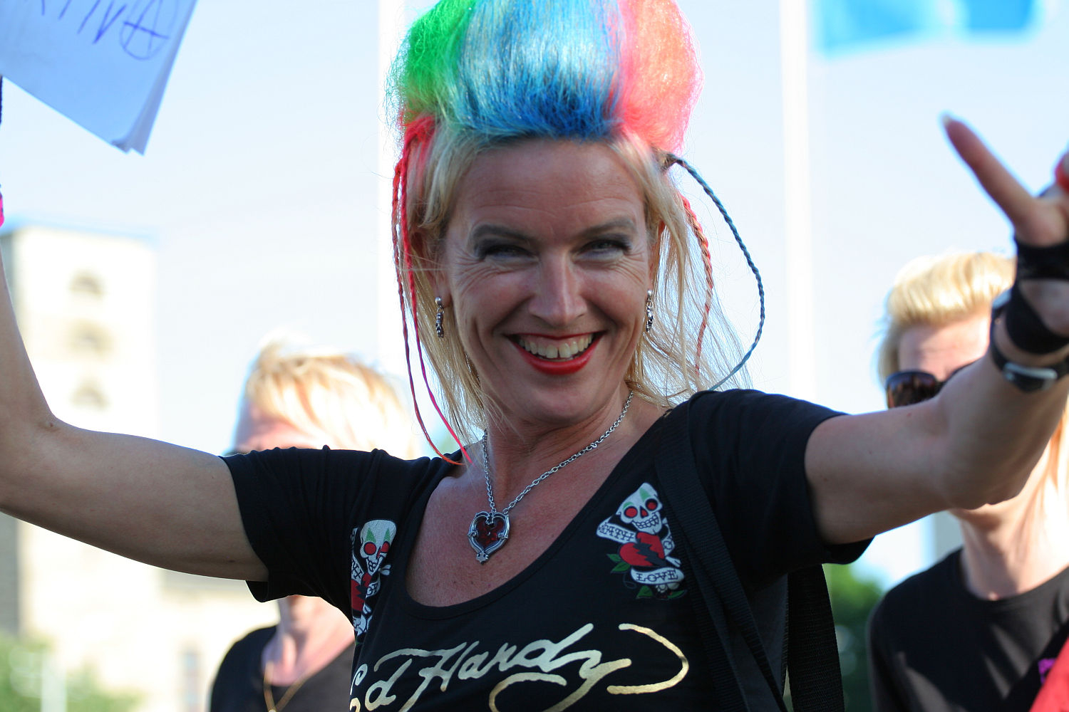 A person with brightly colored, rainbow hair smiles widely. They are wearing a punk-inspired black Ed Hardy shirt with skull designs and a heart-shaped necklace, raising one arm enthusiastically. The background is slightly blurred with blue and white Laulupidu flags and another person partially visible.