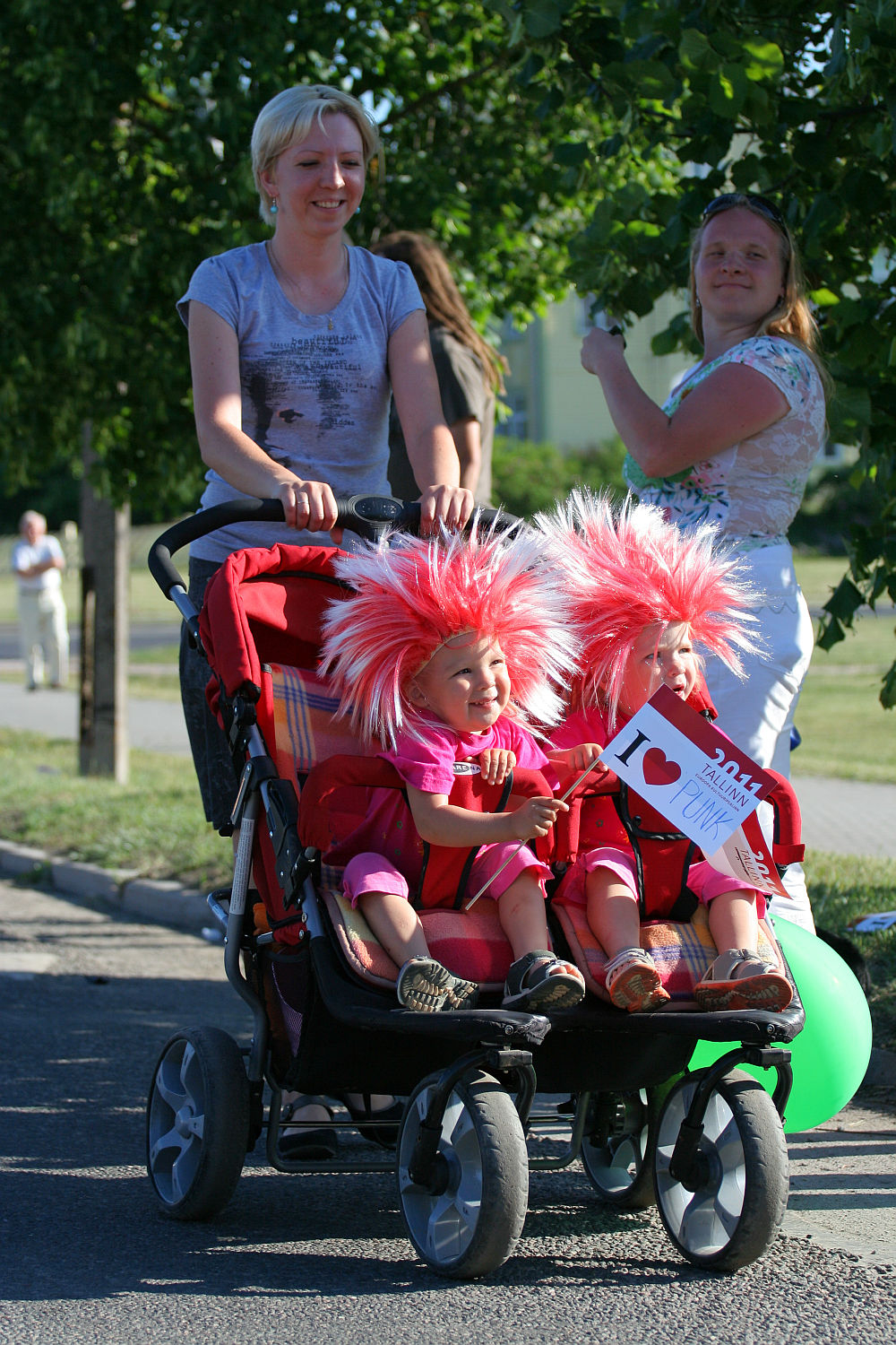 A smiling woman in a punk-inspired outfit is pushing a double stroller with two young children. The children are wearing vibrant red costumes with large, red-and-white striped wigs and holding a sign that reads "I ♥ DAD." Another woman, smiling, stands nearby. They are outdoors on a sunny day with trees in the background.