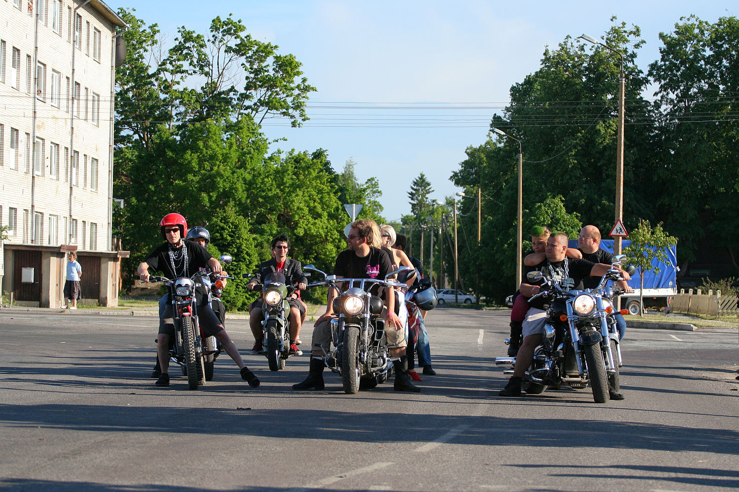 A group of motorcyclists, including one with a red helmet, are lined up on a road in front of a multi-story building and surrounded by tall trees. There are six motorcycles with riders sitting on them, reminiscent of a punk gathering, while people casually gather around or walk in the background.