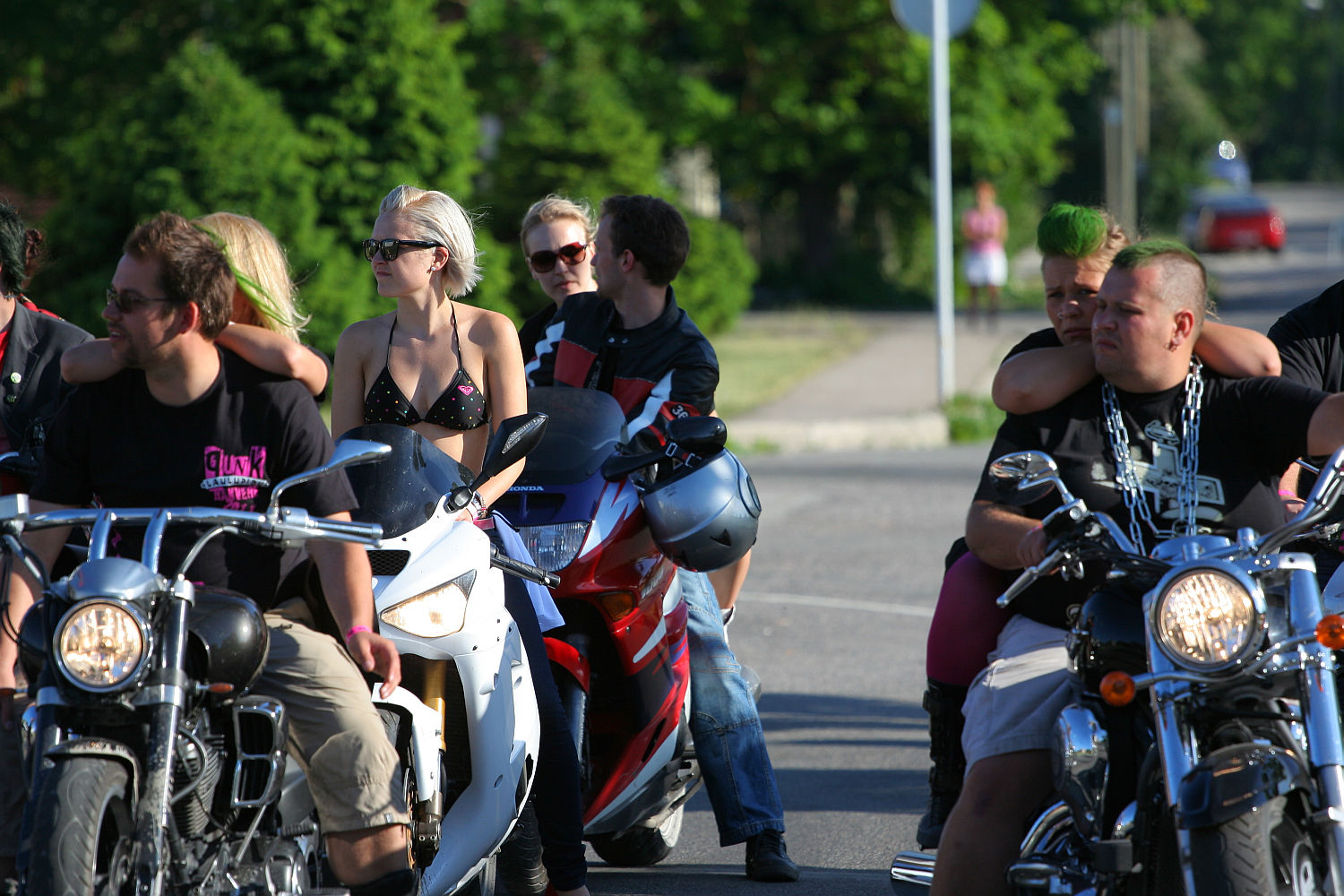 A group of motorcyclists, including men and women, are gathered on the street. Several sport sunglasses and casual clothing, with a distinct punk vibe highlighted by one woman in a black top on a white motorcycle and a man rocking a green mohawk. Trees and blurred street details provide the backdrop.